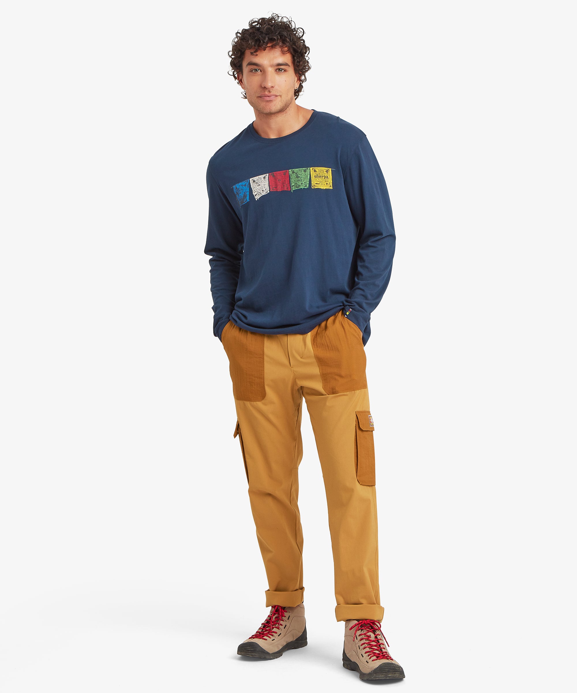 A full-length view of mustard-yellow cargo trousers with ripstop pockets, worn with a navy long-sleeve top printed with multicolored Tarcho flag graphics.