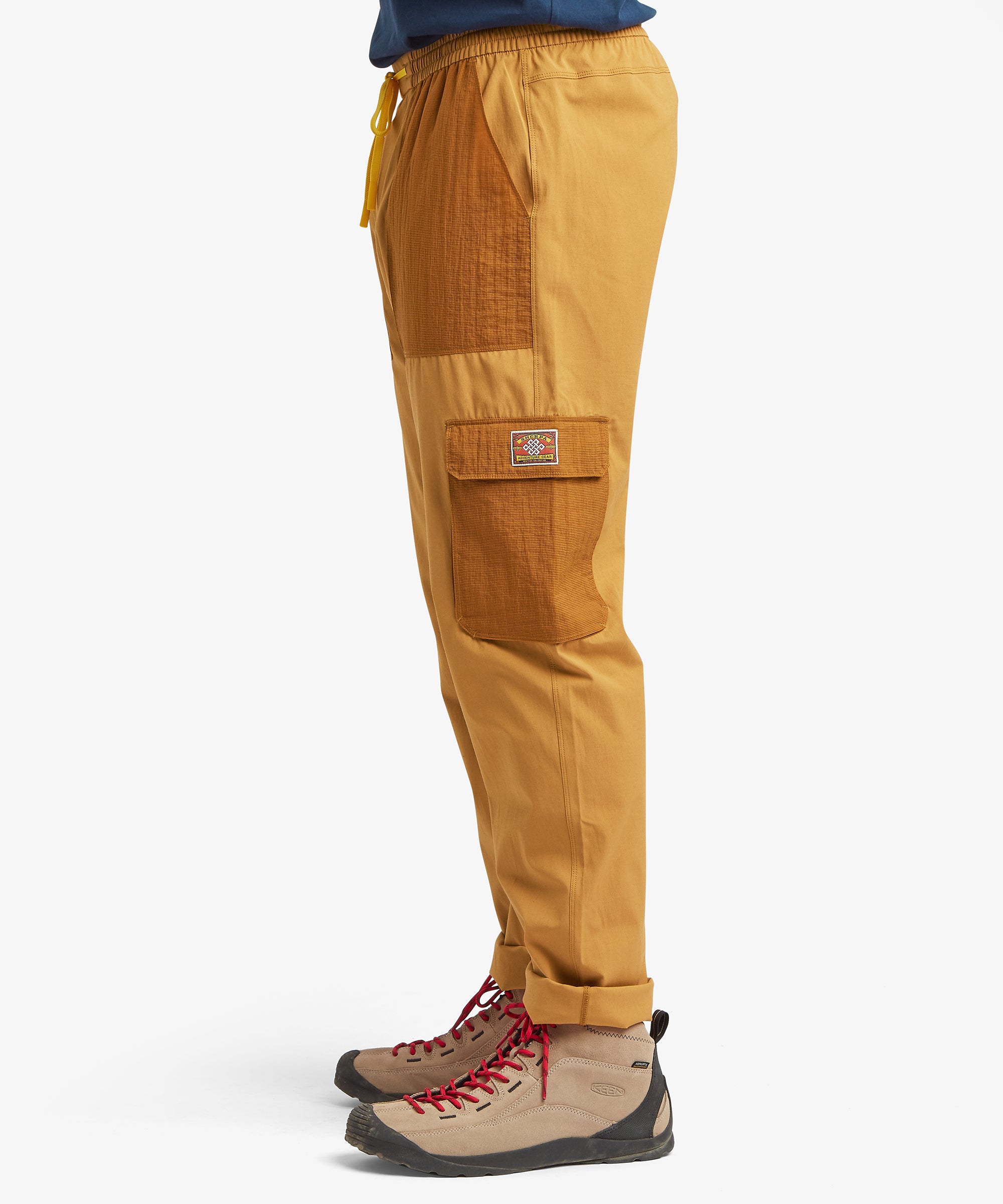 A side view of mustard-yellow outdoor cargo trousers featuring oversized ripstop patch pockets at the thigh and hip, styled with tan hiking boots featuring red laces.
