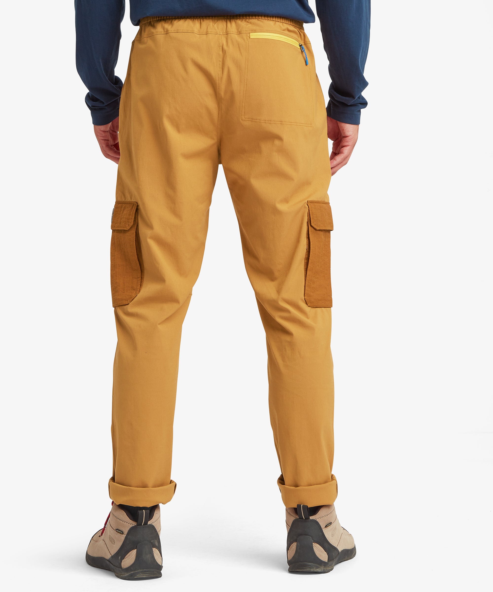 A rear view of mustard-yellow cargo trousers showing dual ripstop cargo pockets and a contrast-trimmed back pocket. Styled with tan hiking boots.