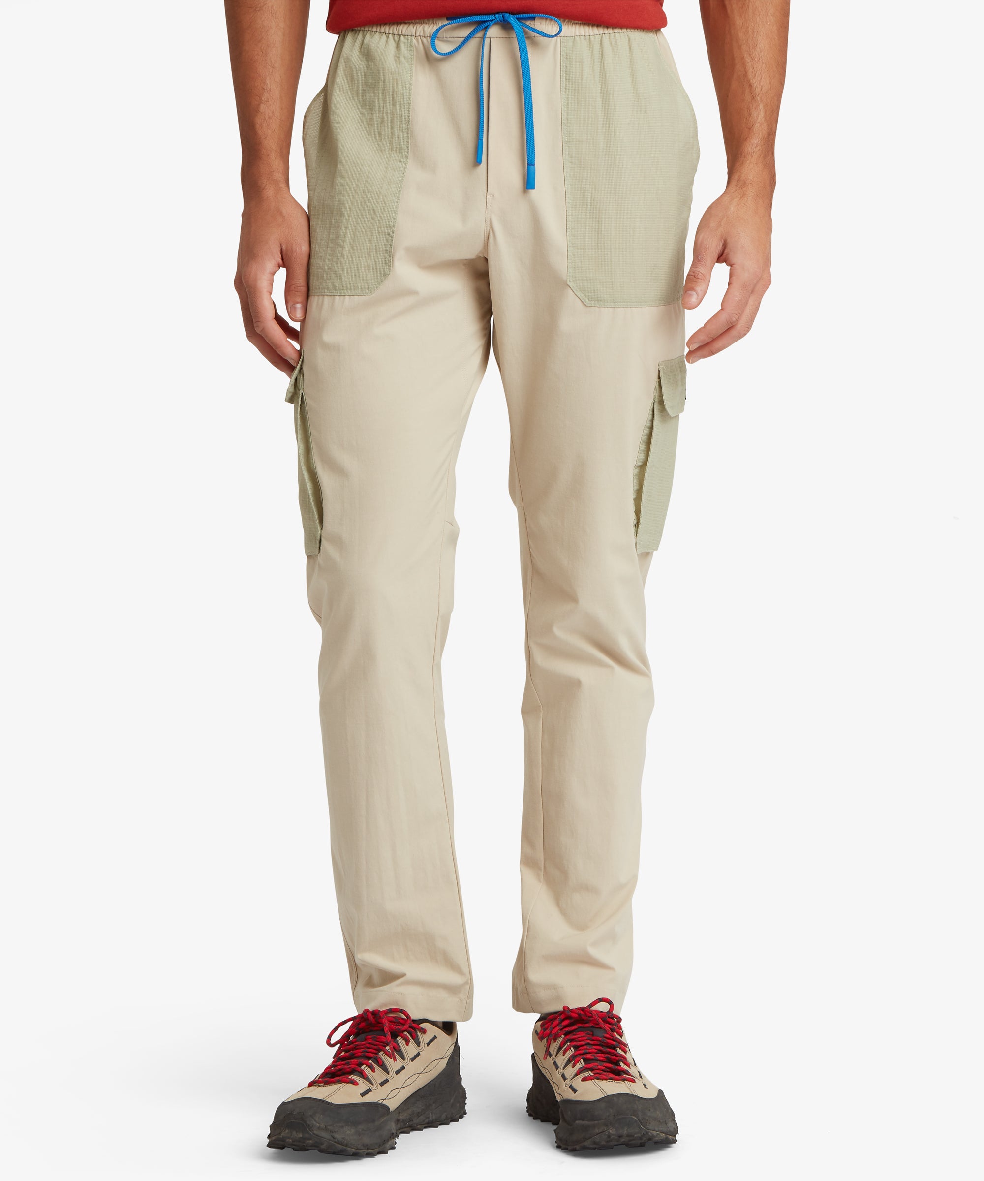 A front view of lightweight beige cargo trousers featuring sage-green ripstop patch pockets at the hips and mid-leg cargo pockets. The trousers have an elastic waist with a bright blue drawcord and are paired with hiking shoes.