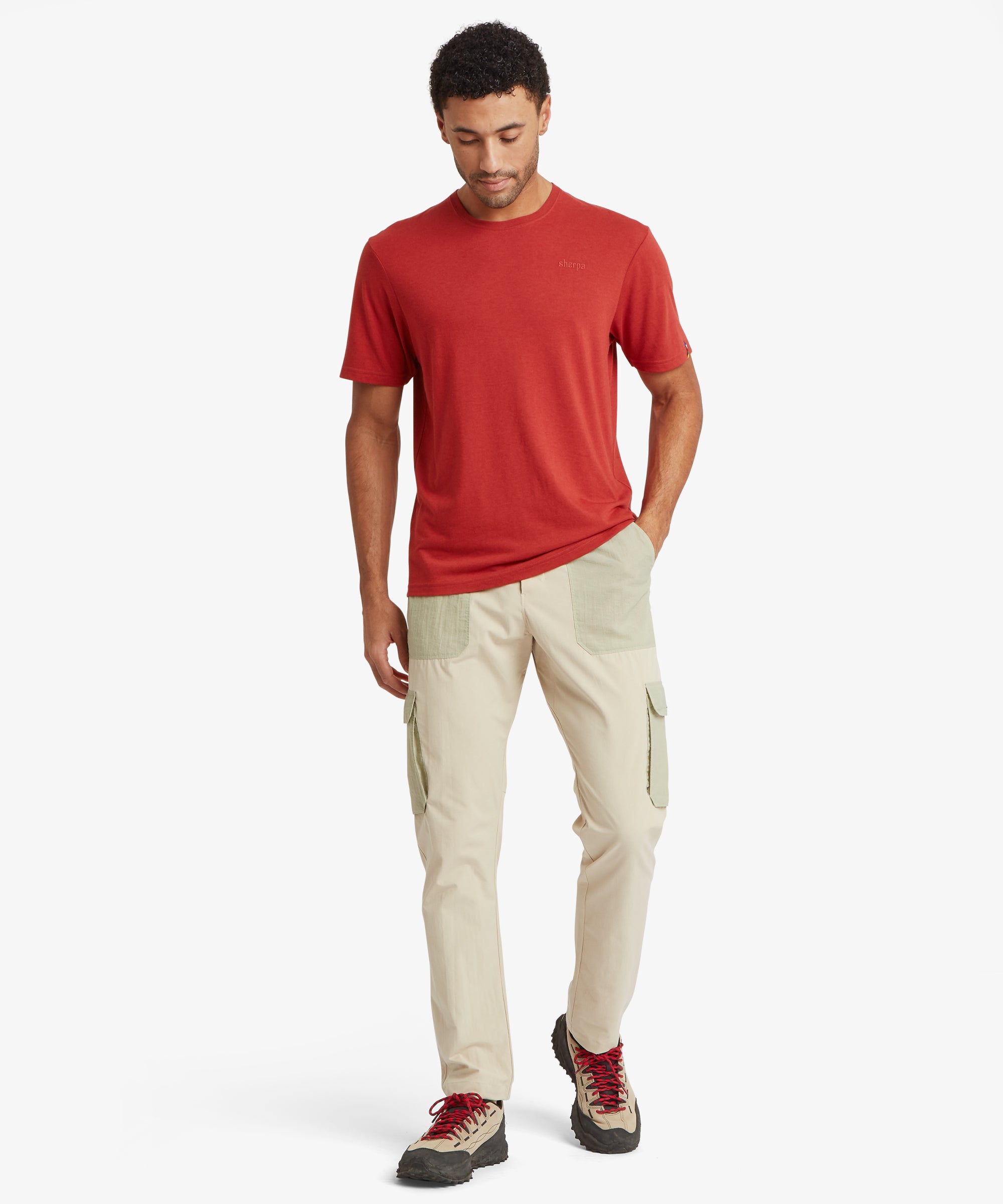 A full-length view of the beige cargo trousers with contrasting sage‑green ripstop pockets at the hips and thighs. Styled with a red short‑sleeve top and outdoor shoes with chunky black soles and red laces.