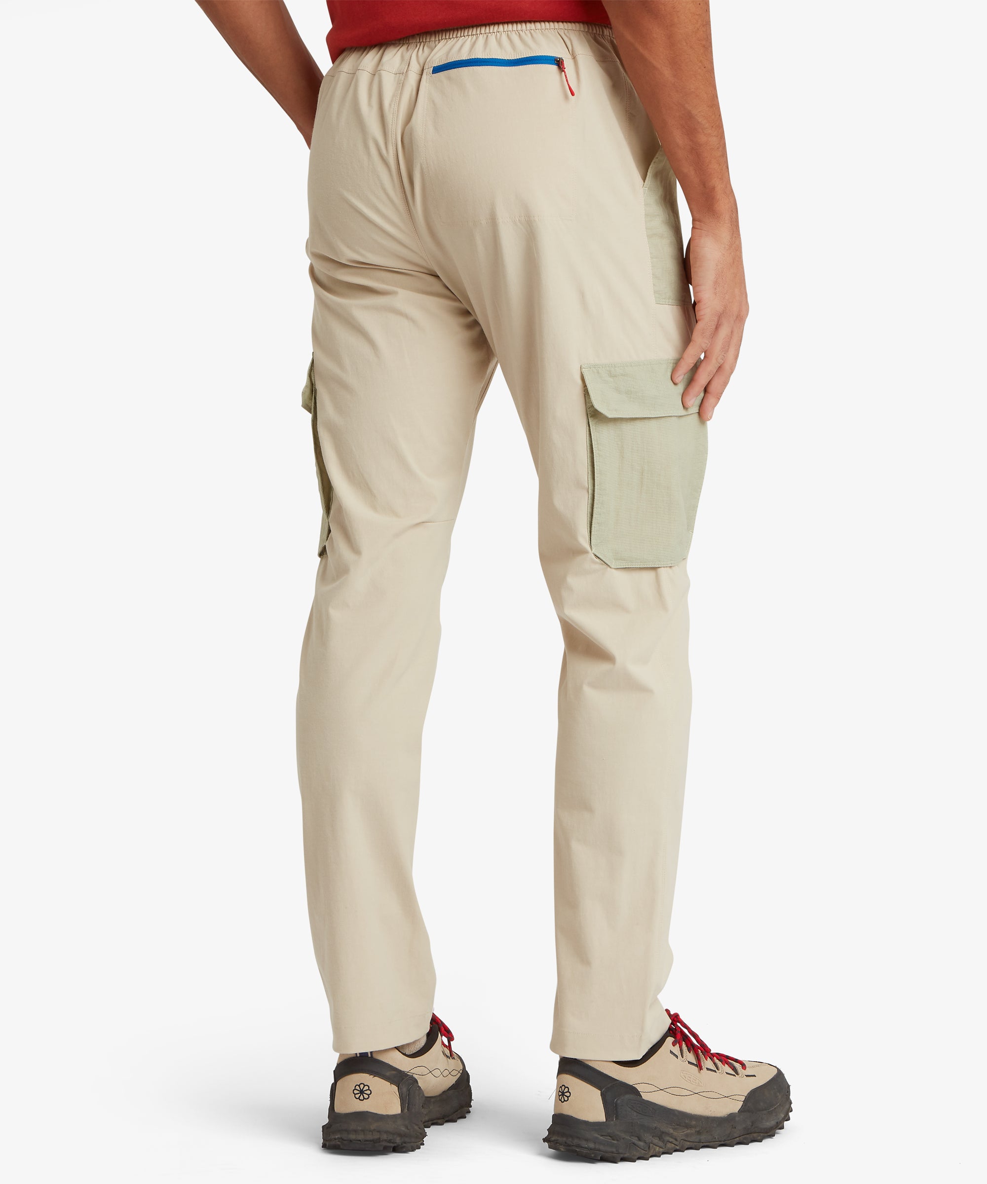 A rear view of the beige cargo trousers showing the elastic waistband, a back pocket with bright blue trim, and cargo pockets on both legs in sage‑green ripstop fabric. The look is styled with sturdy hiking shoes.