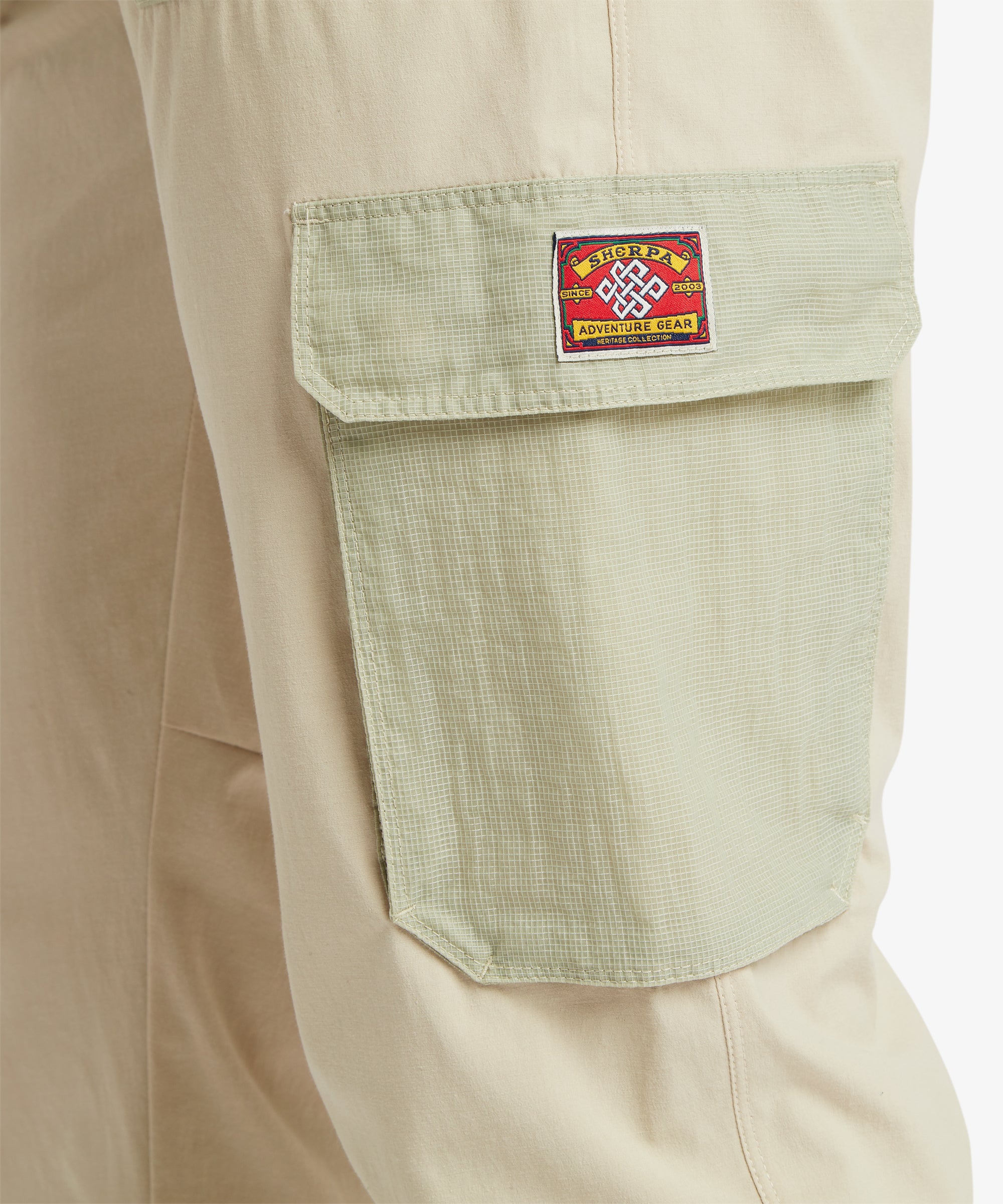 A close-up of the sage‑green cargo pocket on the beige trousers, showing the flap closure and a decorative woven adventure‑gear patch in red, yellow, and green. The ripstop texture and reinforced stitching are clearly visible.