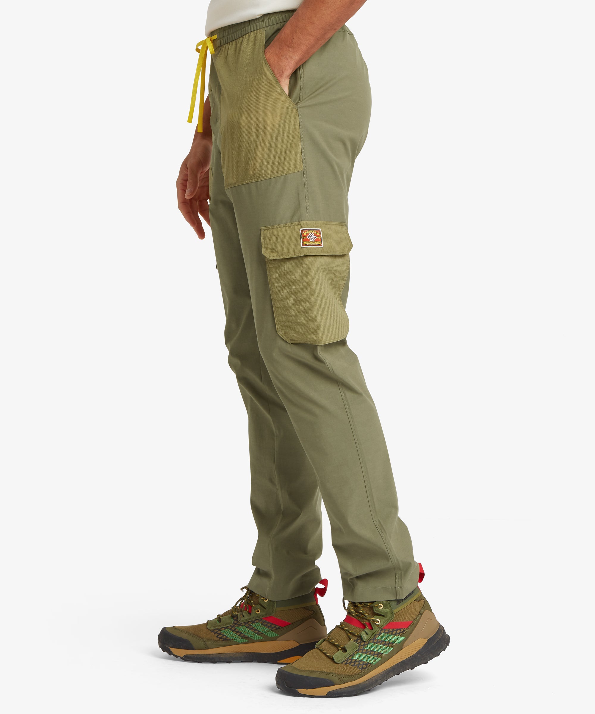 Side view of olive green outdoor trousers showing ripstop utility pockets on the thigh and hip, finished with a bright yellow drawcord at the waistband.