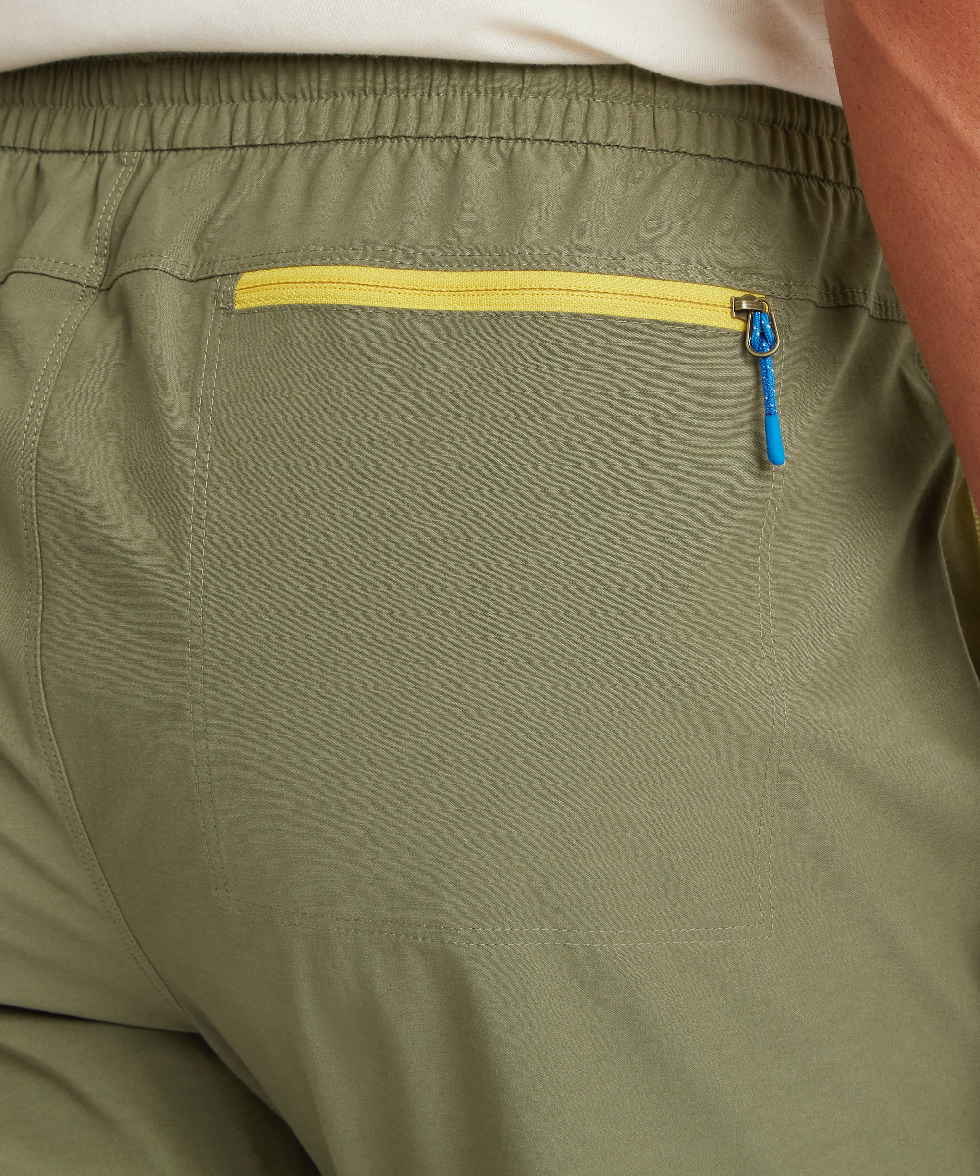 Close‑up of the back pocket on olive outdoor pants, featuring a yellow zipper with a blue pull, combining secure storage with contrast colour detailing.