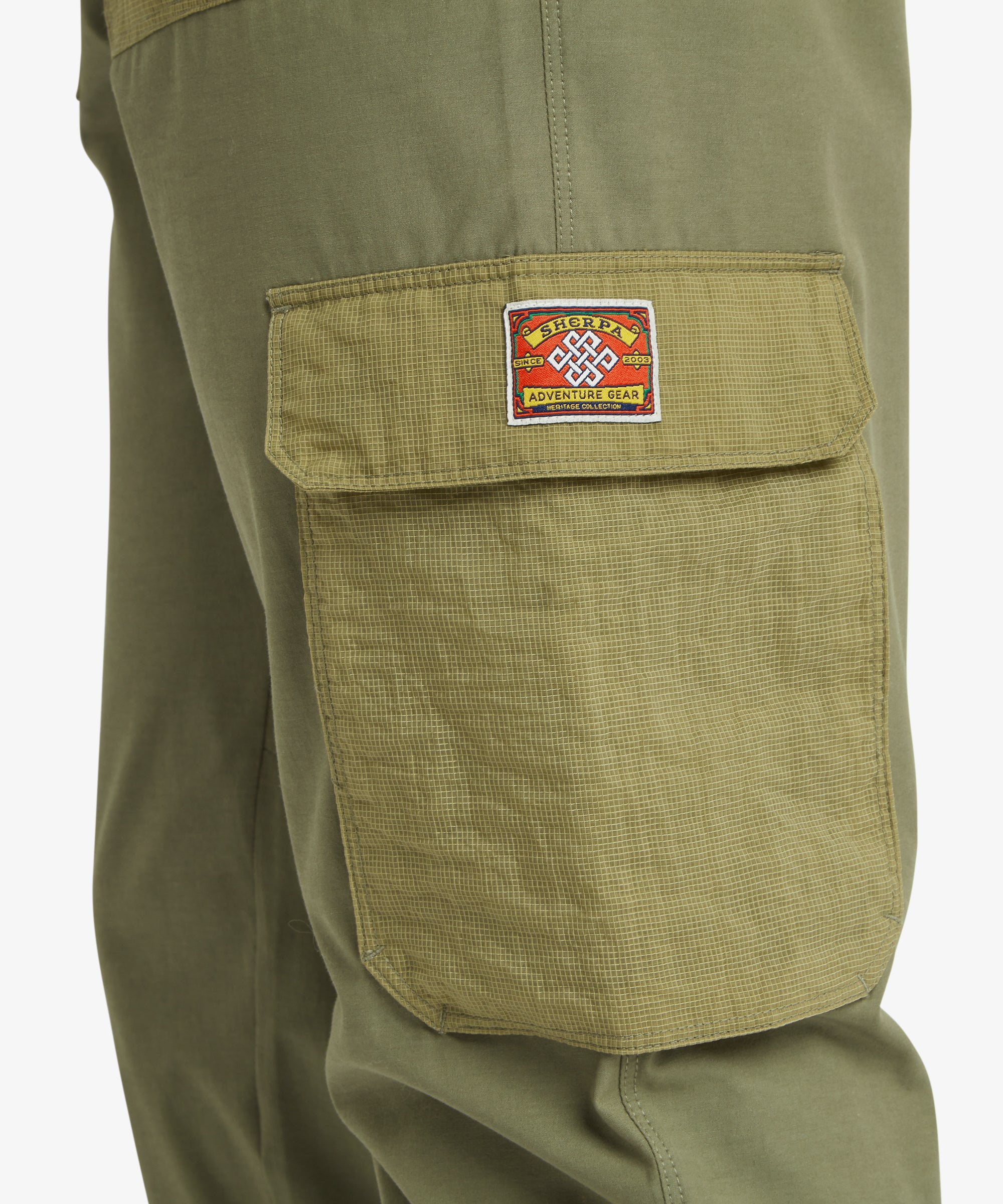 Detail shot of an olive cargo pocket featuring a textured ripstop fabric and a distinctive Sherpa patch, emphasising the pant’s adventure‑ready features.