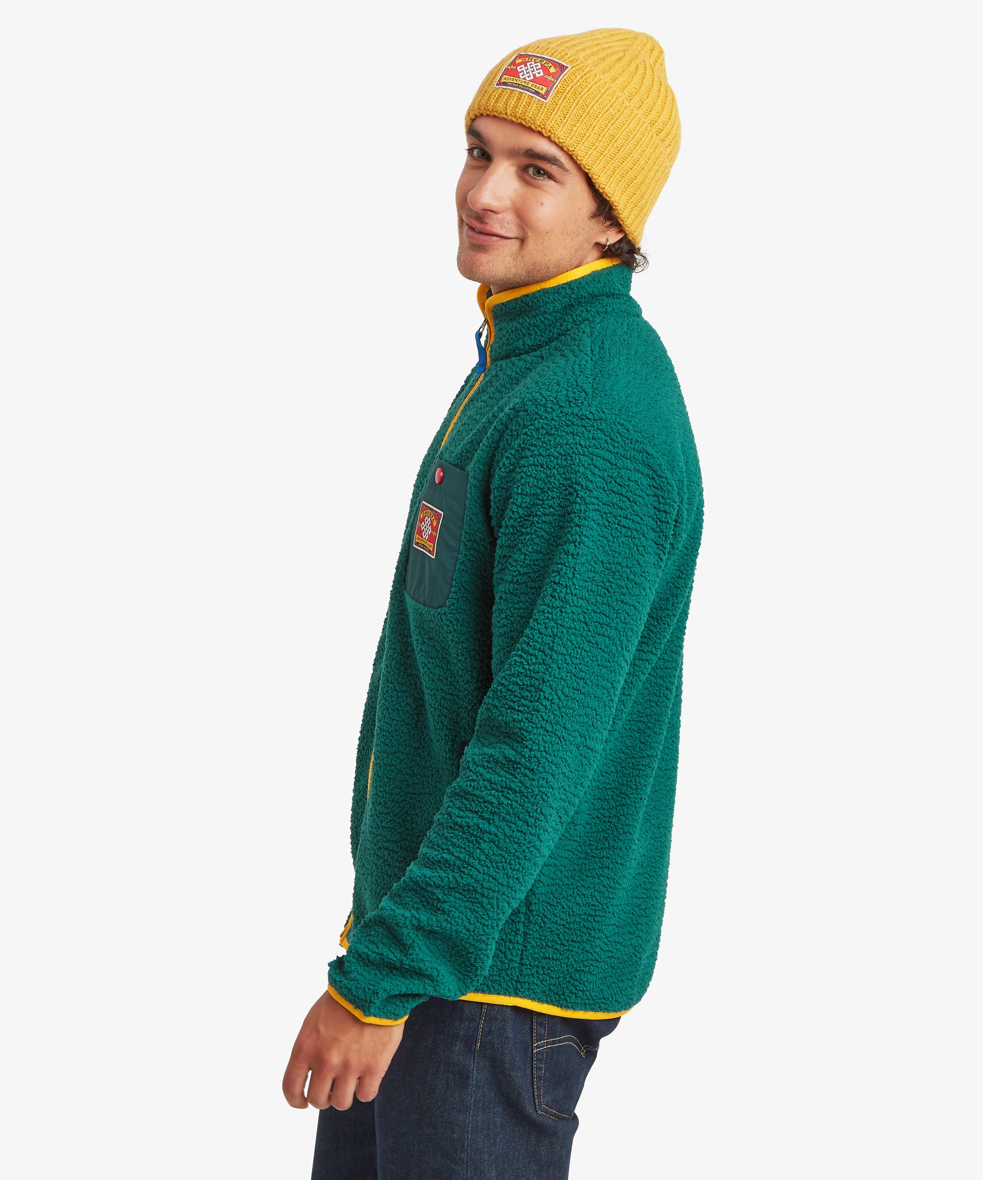 A man wearing a green full zi[ fleece pullover with yellow trim and a yellow knit beanie, shown in profile against a light gray background.