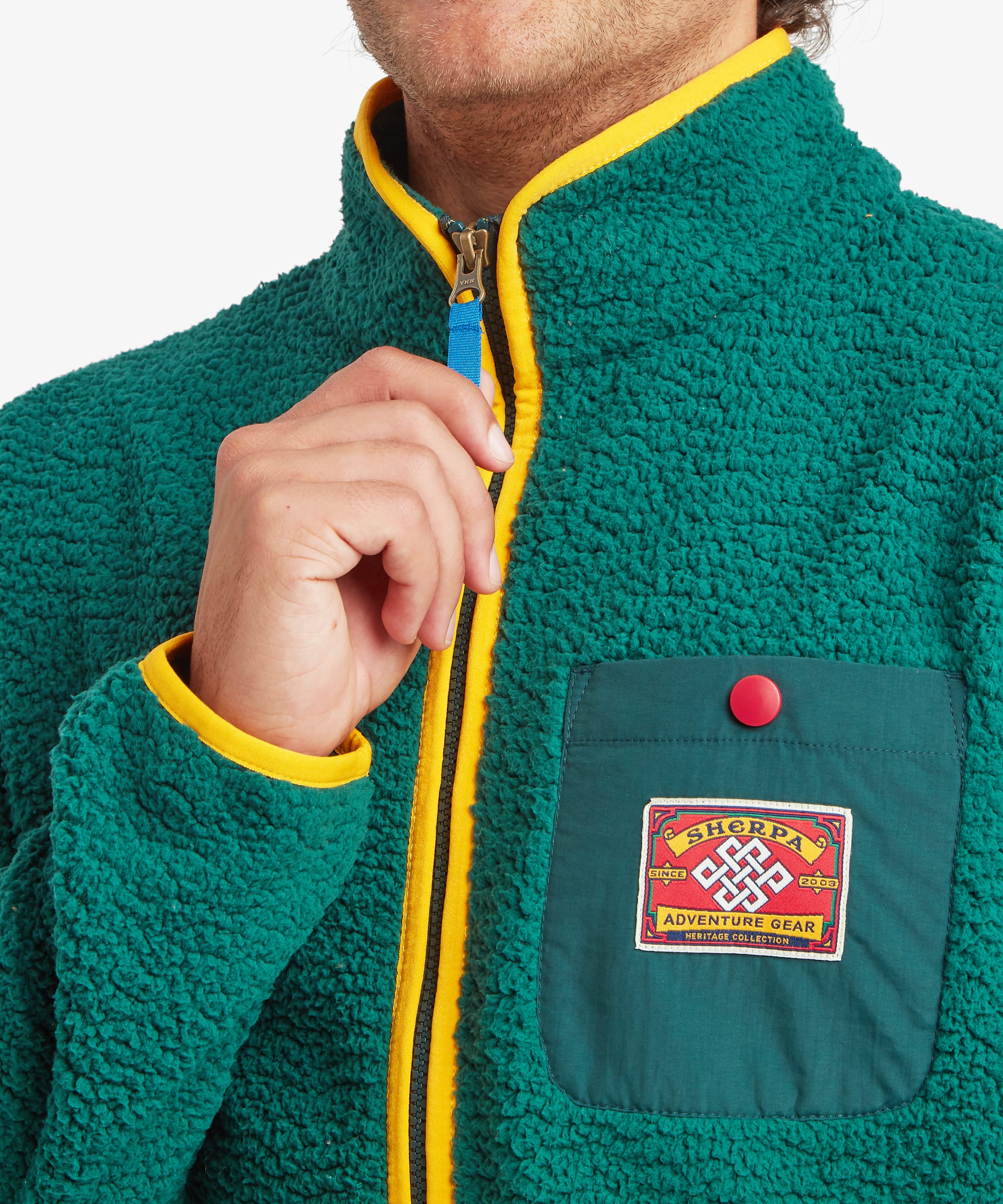 Close-up of a person wearing a green sherpa fleece zip-up jacket with bright yellow trim, featuring a chest pocket with a red snap button and a red 'Sherpa Adventure Gear' patch.