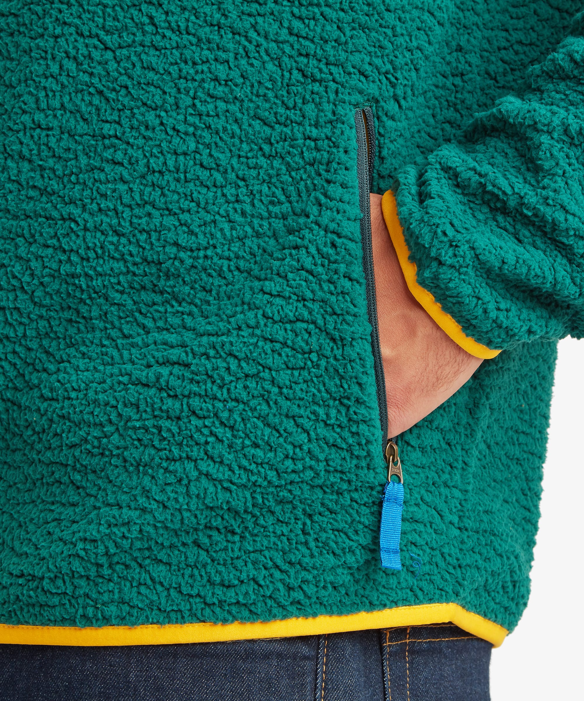 Close-up of a green sherpa fleece zip-up jacket with yellow trim and a blue zipper pull, shown on a person wearing dark blue jeans.