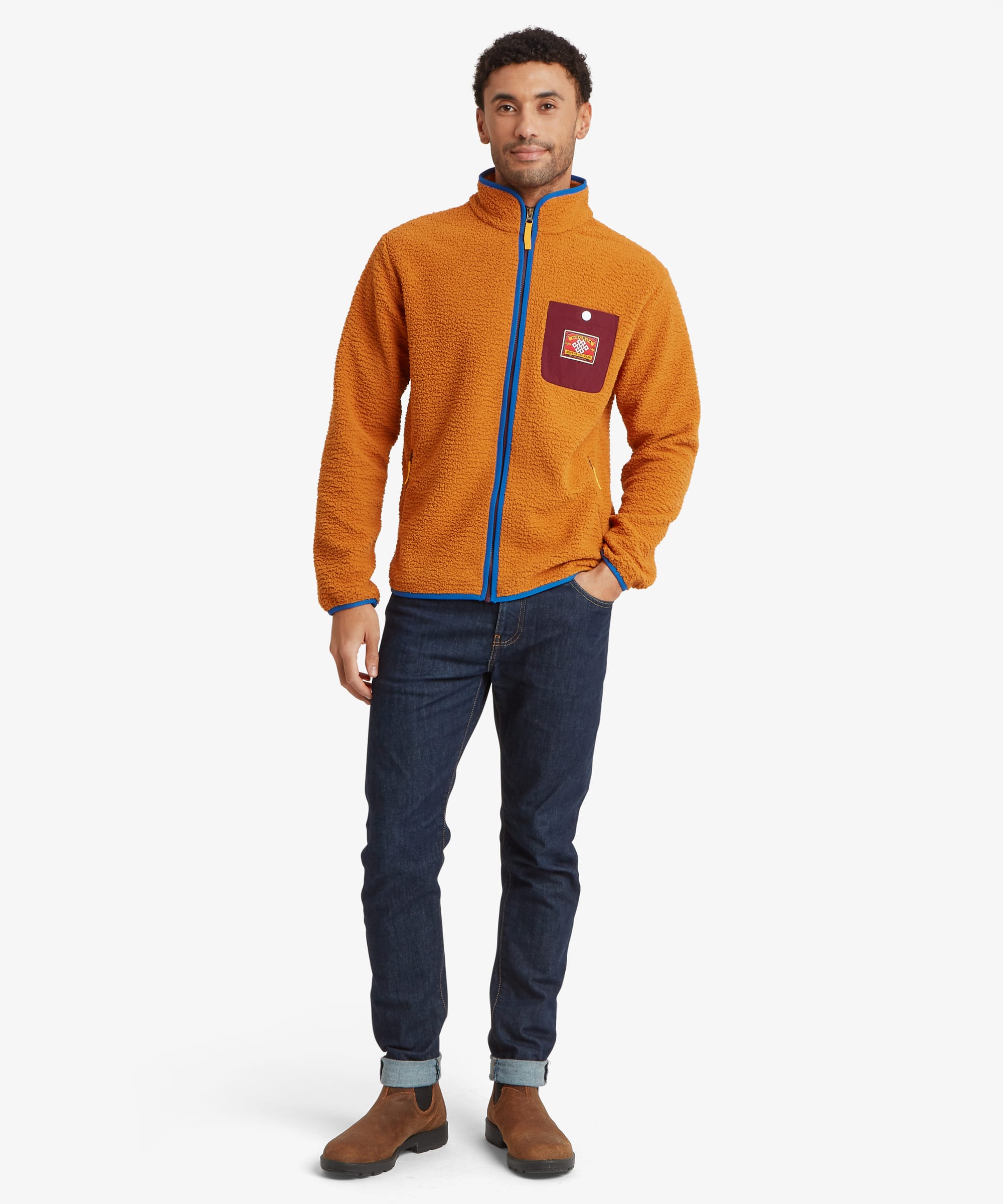 Full-body front view of a model wearing an orange fleece full-zip jacket with blue contrast trim, zippered pockets, and a logo patch on the chest pocket.