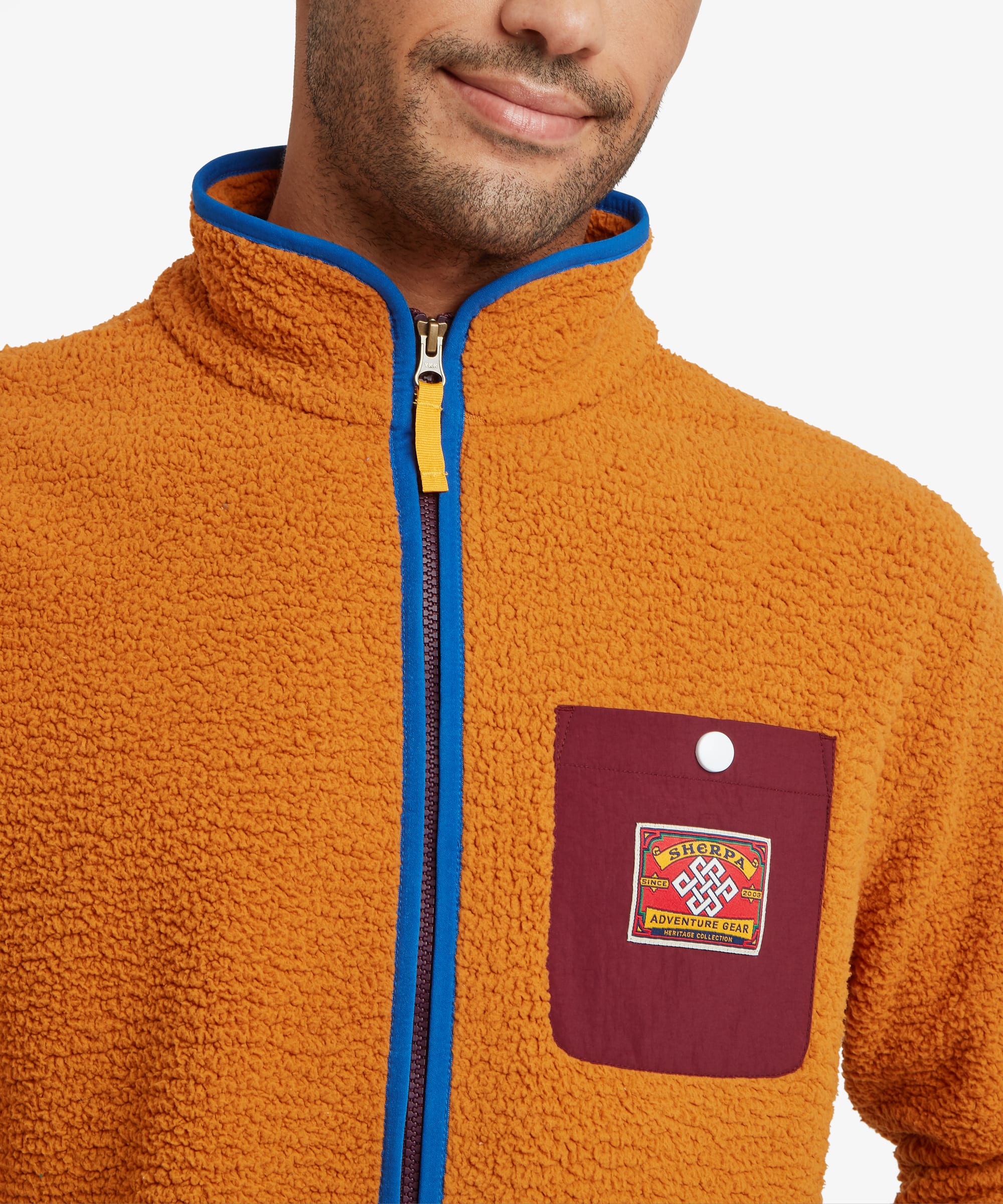 Close-up detail of the orange fleece jacket highlighting the blue front zipper, stand collar, contrast chest pocket, and colorful woven logo patch.