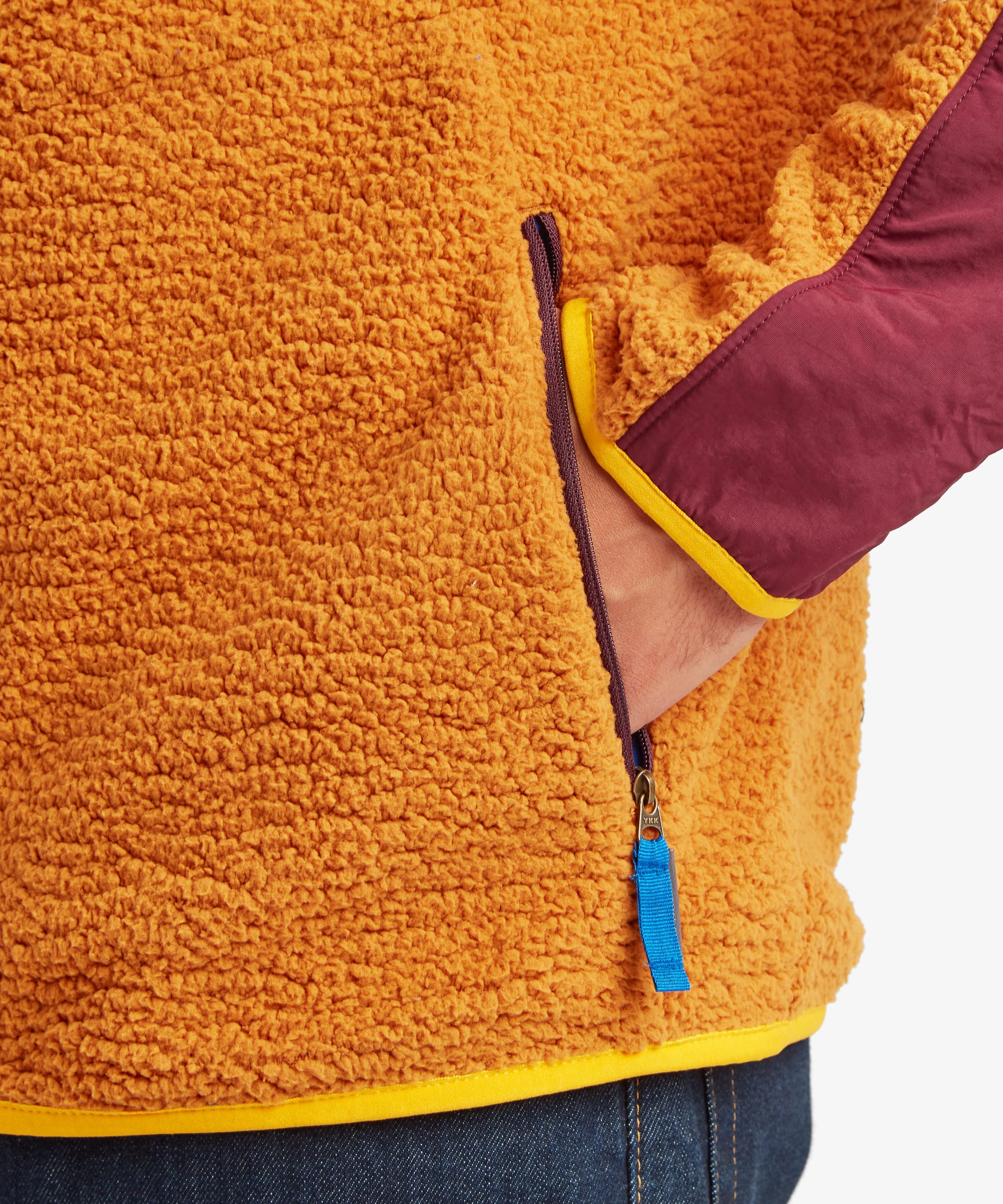 Close-up of the yellow Sherpa Tarcho Hooded Jacket showing the zipped hand pocket, textured fleece fabric and contrasting trim on the hem and cuff. The zip pocket is finished with a blue fabric zip puller.