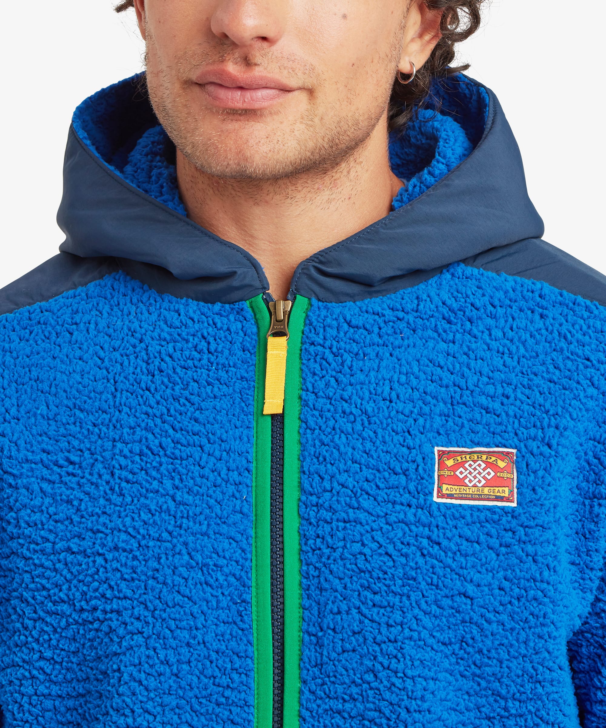 Close-up of a male model wearing the blue Sherpa Tarcho Hooded Jacket, highlighting the soft high-pile fleece, contrasting hood, contrasting full-length zip and embroidered Sherpa logo.