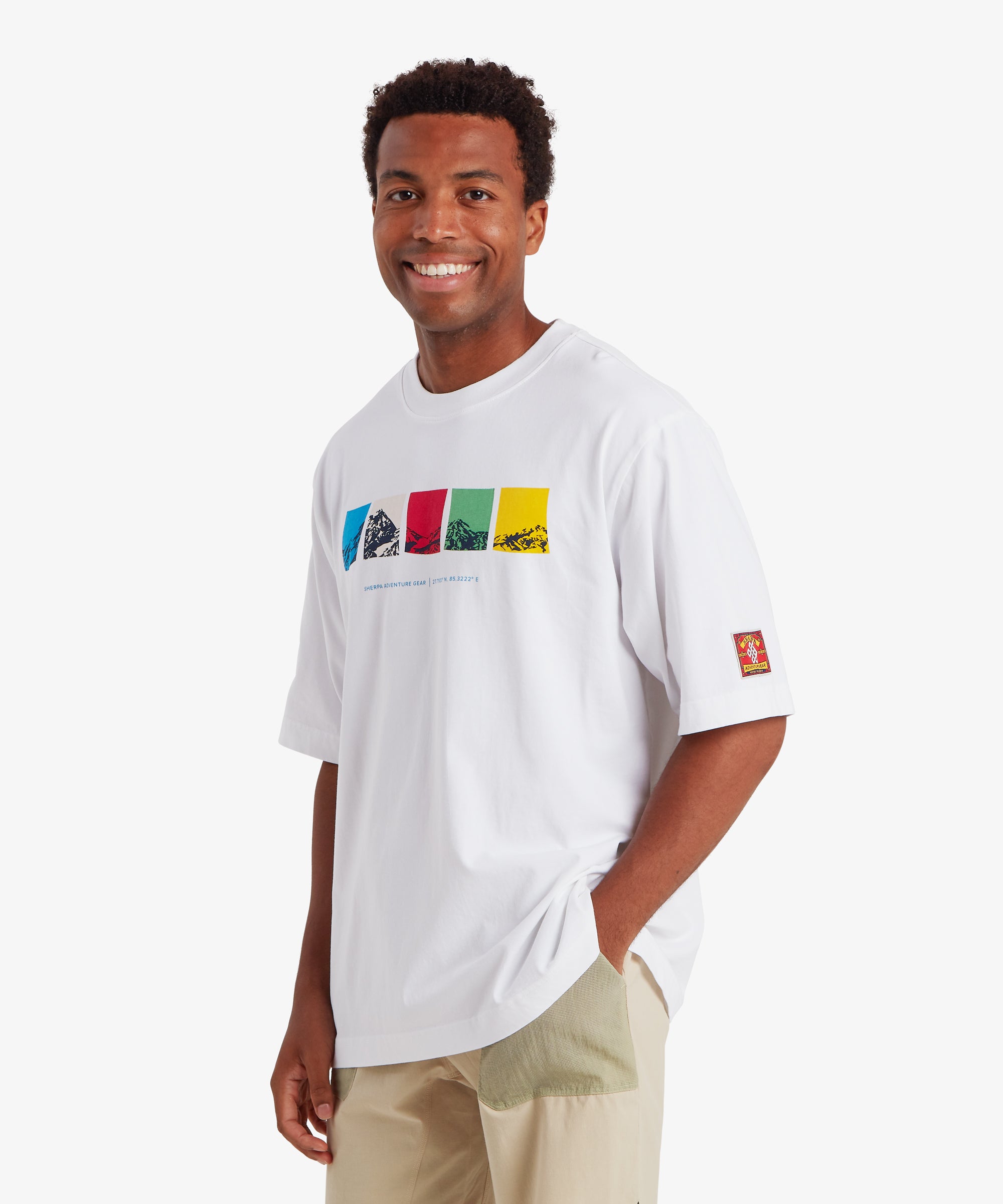 A man wearing a loose fit white T-shirt. The T-shirt features a horizontal graphic across the chest made up of five rectangular color blocks in blue, white, red, green, and yellow, each containing a mountain illustration. Beneath the graphic the words "Sherpa Adventure Gear" are printed in a light blue font. On the sleeve is red, rectangular mountaineering patch.  The outfit is completed with a pair of beige shorts with pale green pockets. The man is standing facing forward with his left hand in his pocket.