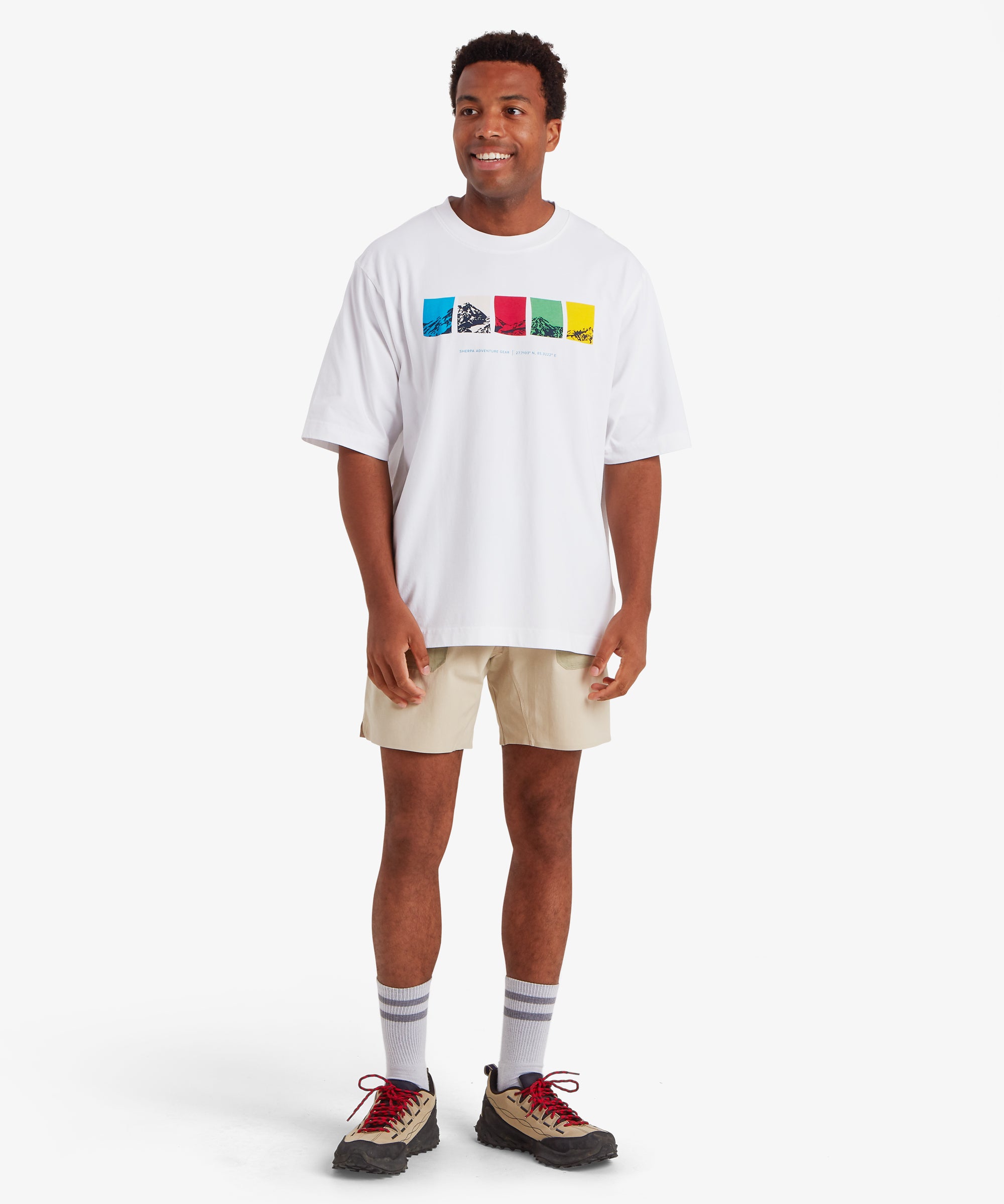 A man wearing a relaxed fit white T-shirt. The T-shirt features a horizontal graphic across the chest made up of five rectangular color blocks in blue, white, red, green, and yellow, each containing a mountain illustration. Beneath the graphic the words "Sherpa Adventure Gear" are printed in a light blue font.The outfit is completed with a pair of beige shorts, white socks and beige shoes with red shoe laces. The man is standing with his body facing forward and his head turned slightly to right.