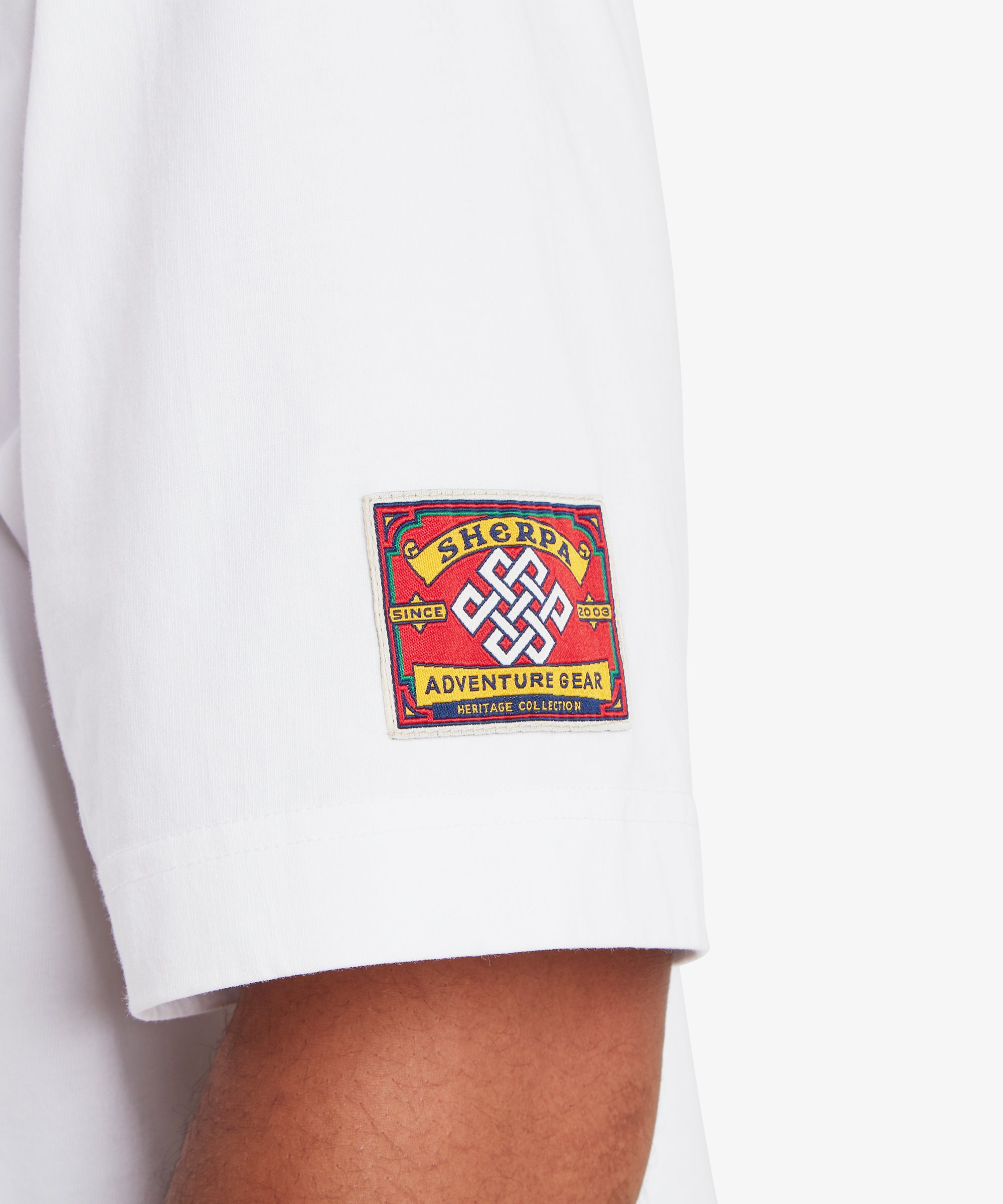 A close-up view of the left sleeve of a white T-shirt. A square red woven patch is stitched near the sleeve hem, featuring a white endless knot symbol at the center. The patch reads “Sherpa Adventure Gear” with “Since 2003” and “Heritage Collection” incorporated into the design. The fabric appears soft with a relaxed drape, set against a white background.
