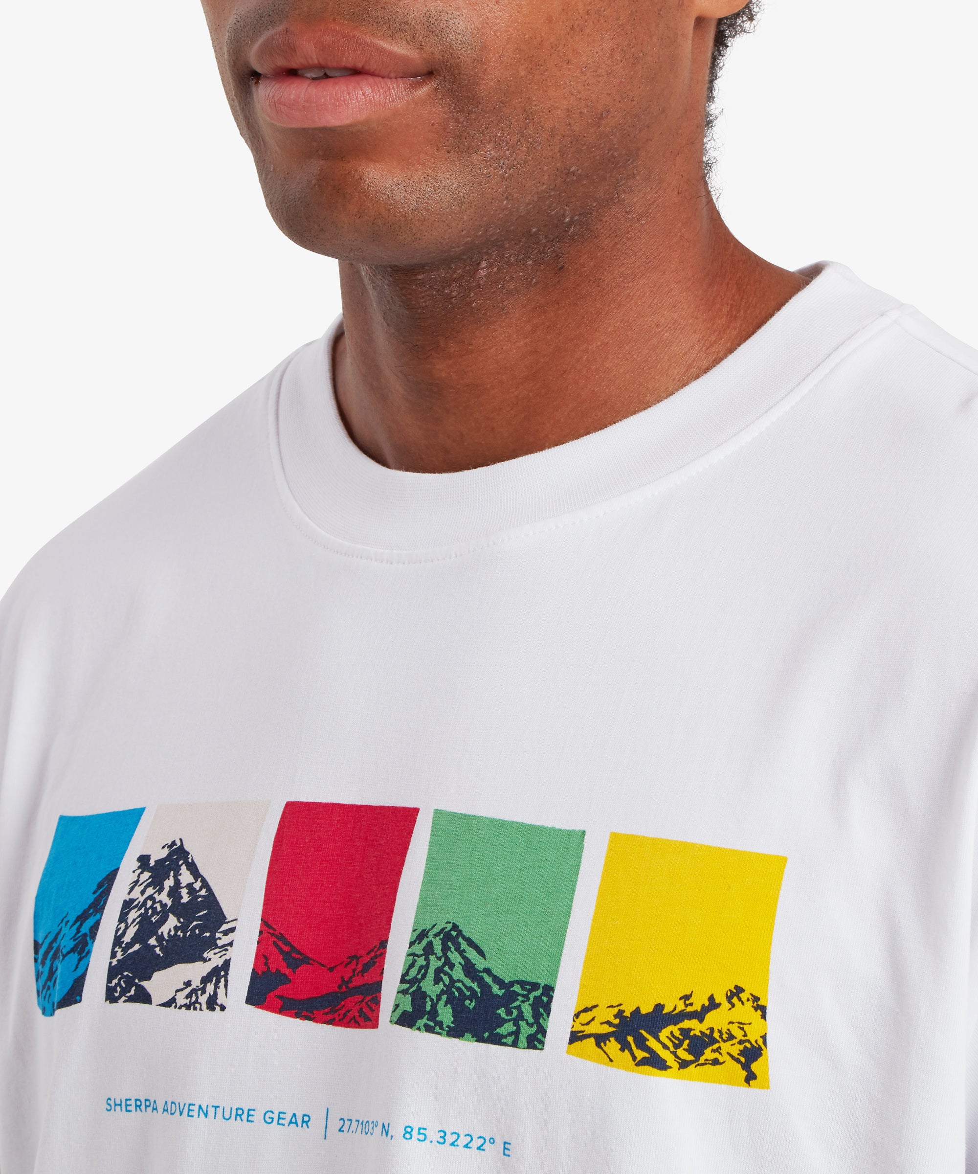 A close-up view of a man wearing a white T-shirt. The man's upper chest and the neckline of the T-shirt are visible. The T-shirt has a ribbed crew neck collar and features a horizontal graphic across the chest made up of five rectangular color blocks in blue, white, red, green, and yellow, each containing a stylized mountain illustration. Beneath the graphic is small text reading “Sherpa Adventure Gear” followed by geographic coordinates. The man is standing against a white background.