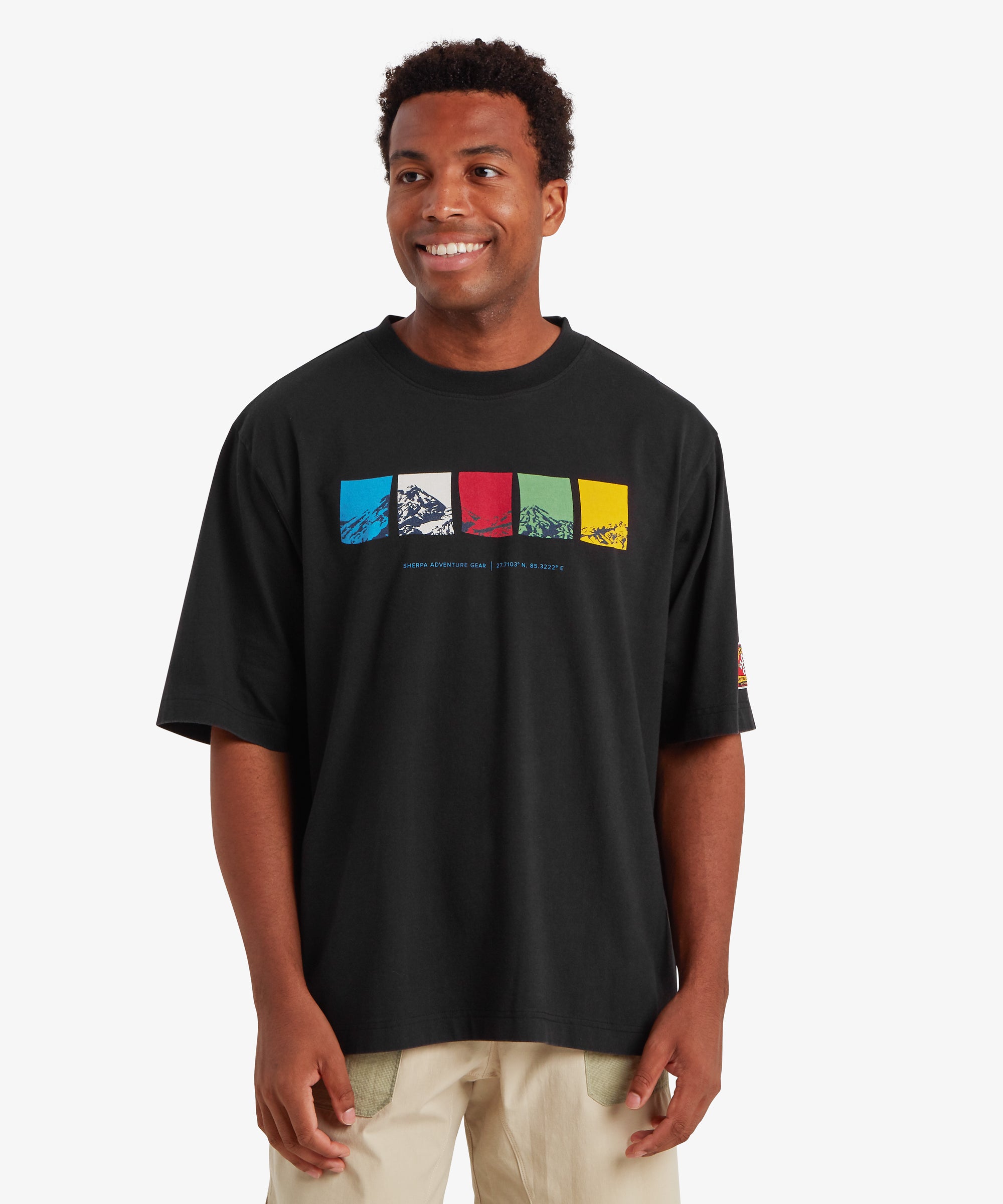 A man wearing a relaxed fit black T-shirt. The T-shirt features a horizontal graphic across the chest made up of five rectangular color blocks in blue, white, red, green, and yellow, each containing a different mountain illustration. Beneath the graphic the words "Sherpa Adventure Gear" and a set of co-ordinates are printed in a light blue font. The outfit is completed with a pair of beige shorts with pale green pockets. The man is standing facing forward against a white background.