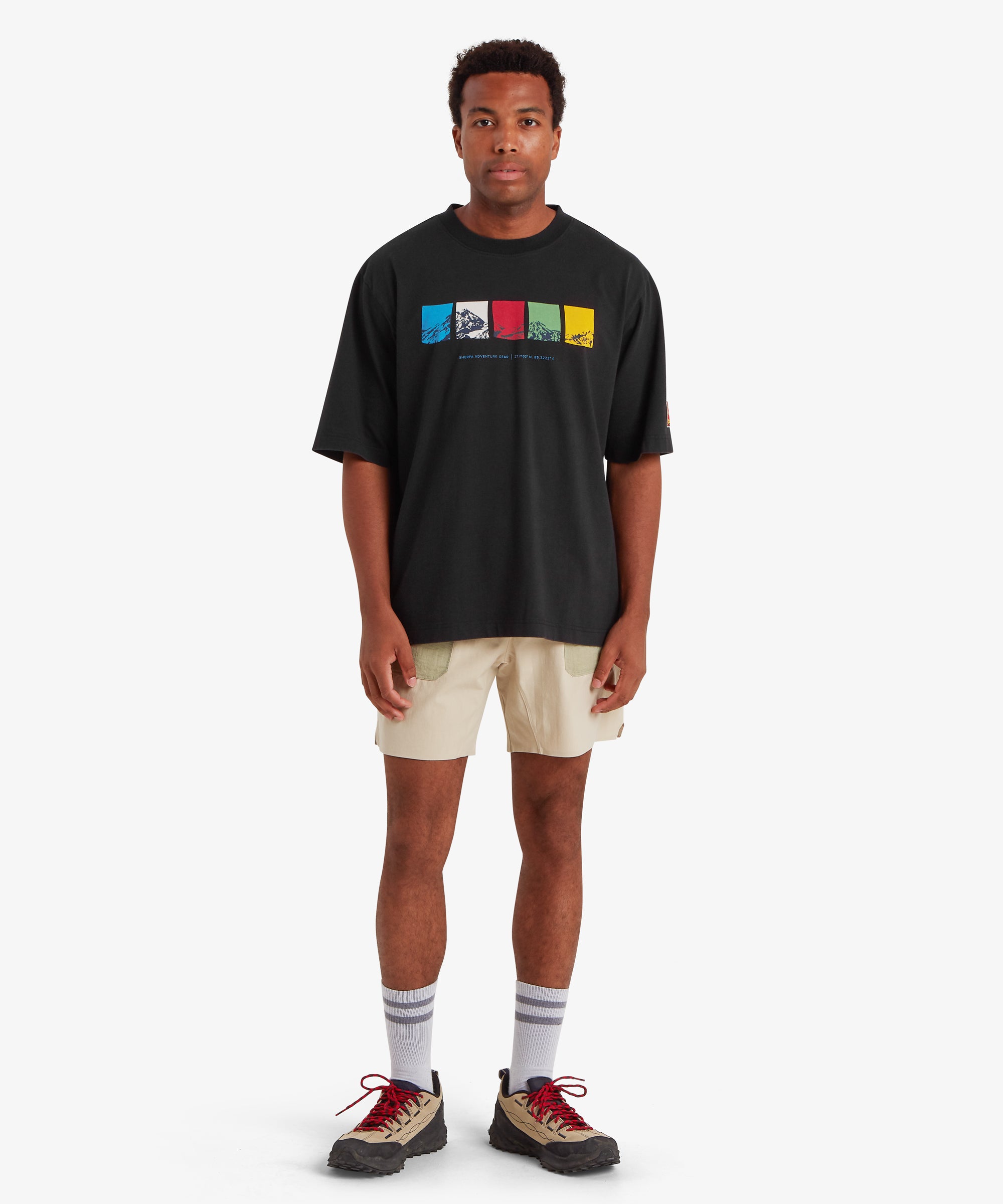 Man wearing a relaxed fit black T-shirt. The T-shirt features a horizontal graphic across the chest made up of five rectangular color blocks in blue, white, red, green, and yellow, each containing a mountain illustration. Beneath the graphic the words "Sherpa Adventure Gear" are printed in a light blue font. On the left sleeve is red, rectangular mountaineering patch.  The outfit is completed with a pair of beige shorts, white socks and beige walking shoes with red laces. The man is facing forwards.