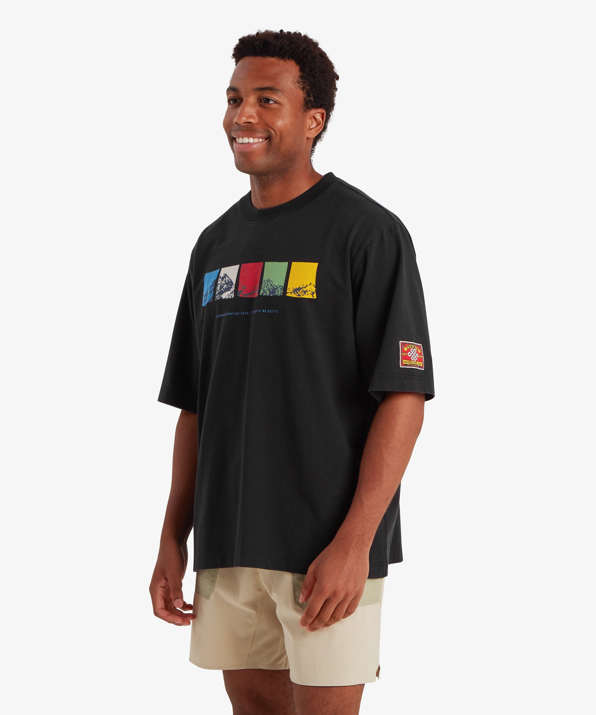 Man wearing a relaxed fit black T-shirt. The T-shirt features a horizontal graphic across the chest made up of five rectangular color blocks in blue, white, red, green, and yellow, each containing a mountain illustration. Beneath the graphic the words "Sherpa Adventure Gear" are printed in a light blue font. On the left sleeve is red, rectangular mountaineering patch.  The outfit is completed with a pair of beige shorts with pale green pockets. The man is standing with is body turned slightly to the right.