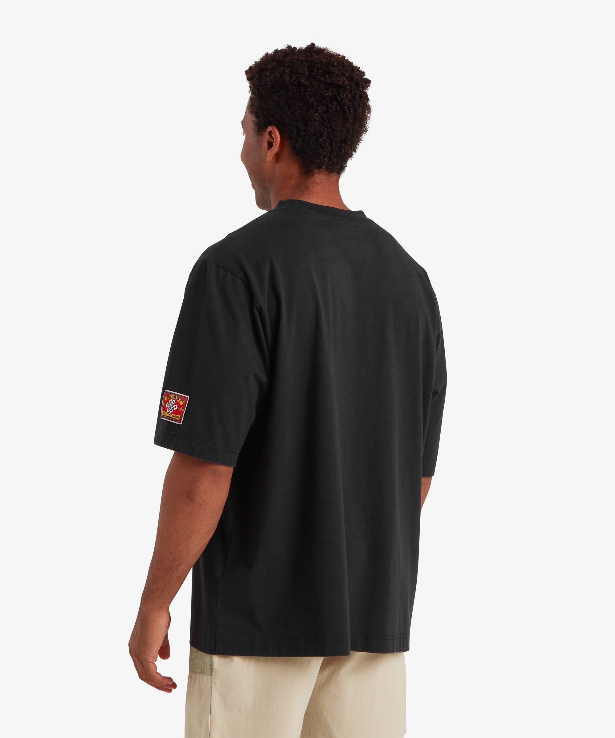 A back view of a man wearing relaxed fit, baggy black T-shirt. The back of T-shirt is plain of the man with no graphic or detail. On the left sleeve is a rectangular mountaineering patch. At the centre of the patch is a white endless knot graphic. The words "Sherpa adventure Gear" are stitched above and below the endless knot. The T-shirt is paired with beige shorts.