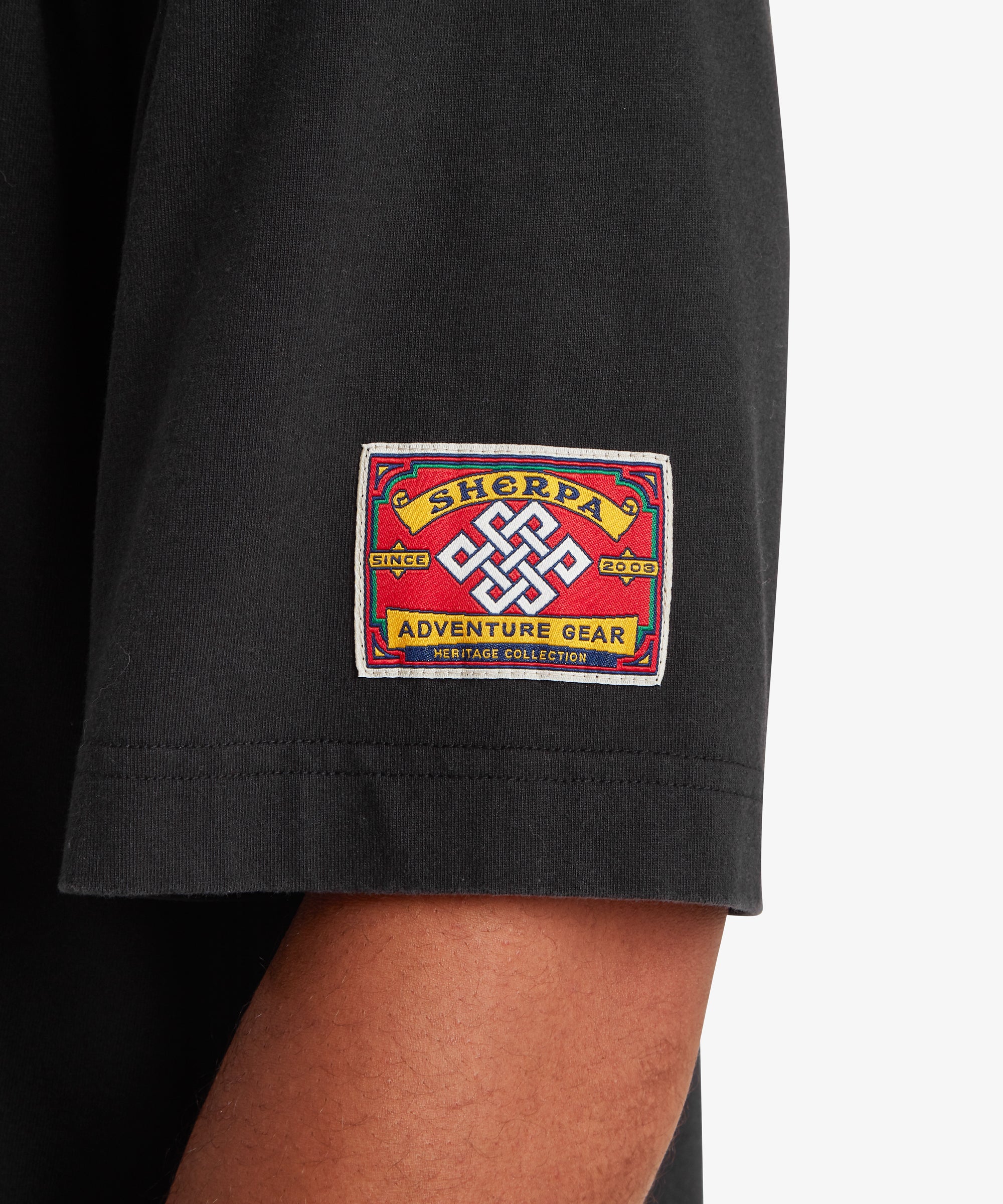 A close-up view of the left sleeve of a black T-shirt. A square red woven patch is stitched near the sleeve hem, featuring a white endless knot symbol at the center. The patch reads “Sherpa Adventure Gear” with “Since 2003” and “Heritage Collection” incorporated into the design. The fabric appears soft with a relaxed drape, set against a white background.