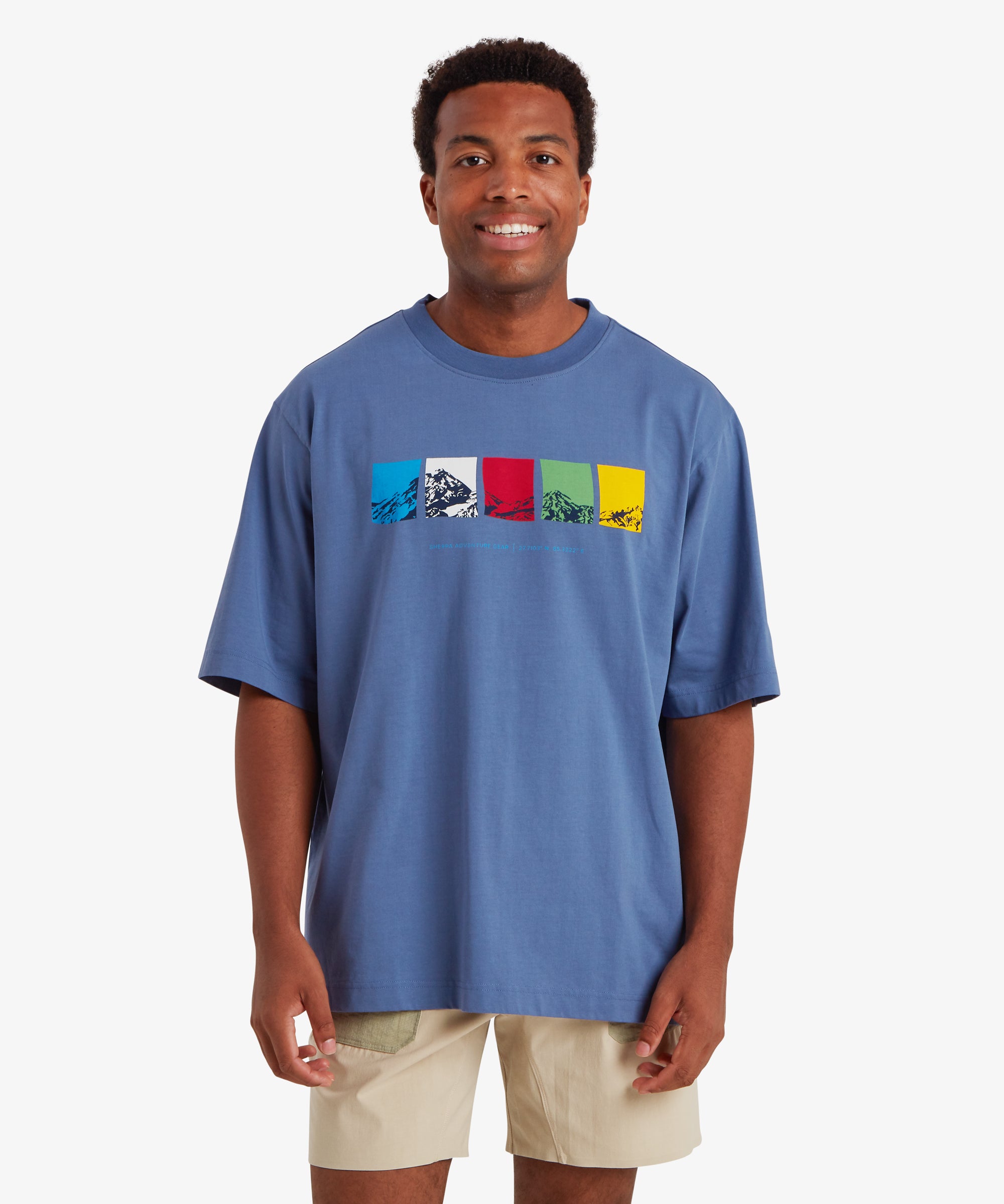 A man wearing a loose, relaxed fit blue T-shirt. The T-shirt features a horizontal graphic across the chest made up of five rectangular color blocks in blue, white, red, green, and yellow, each containing a mountain illustration. Beneath the graphic the words "Sherpa Adventure Gear" and a set of co-ordinates are printed in a light blue font. The outfit is completed with a pair of beige shorts with pale green pockets. The man is standing facing forward against a white background.