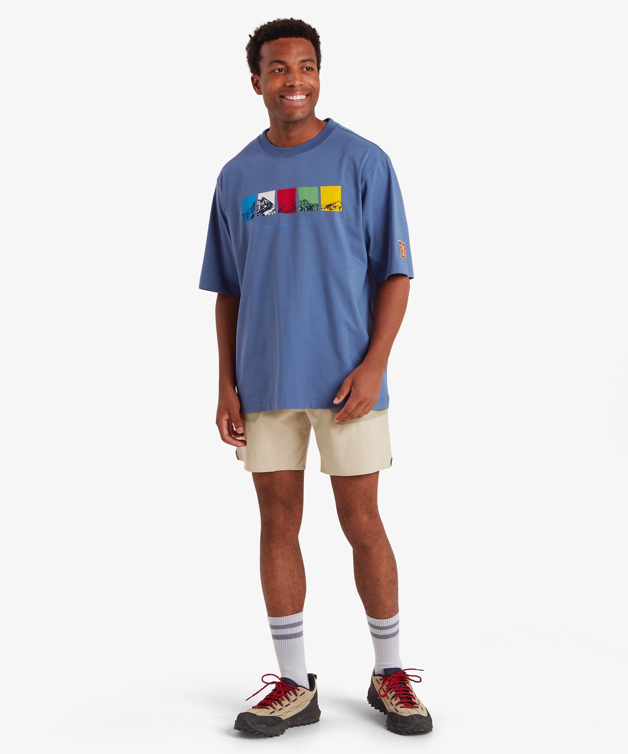 Man wearing a relaxed fit blue T-shirt. The T-shirt features a horizontal graphic across the chest made up of five rectangular color blocks in blue, white, red, green, and yellow, each containing a mountain illustration. Below the graphic the words "Sherpa Adventure Gear" are printed in a light blue font. On the left sleeve is red, rectangular mountaineering patch. The outfit is styled with of beige shorts, white socks and beige walking shoes with red laces. The man has his head turned slightly to the left