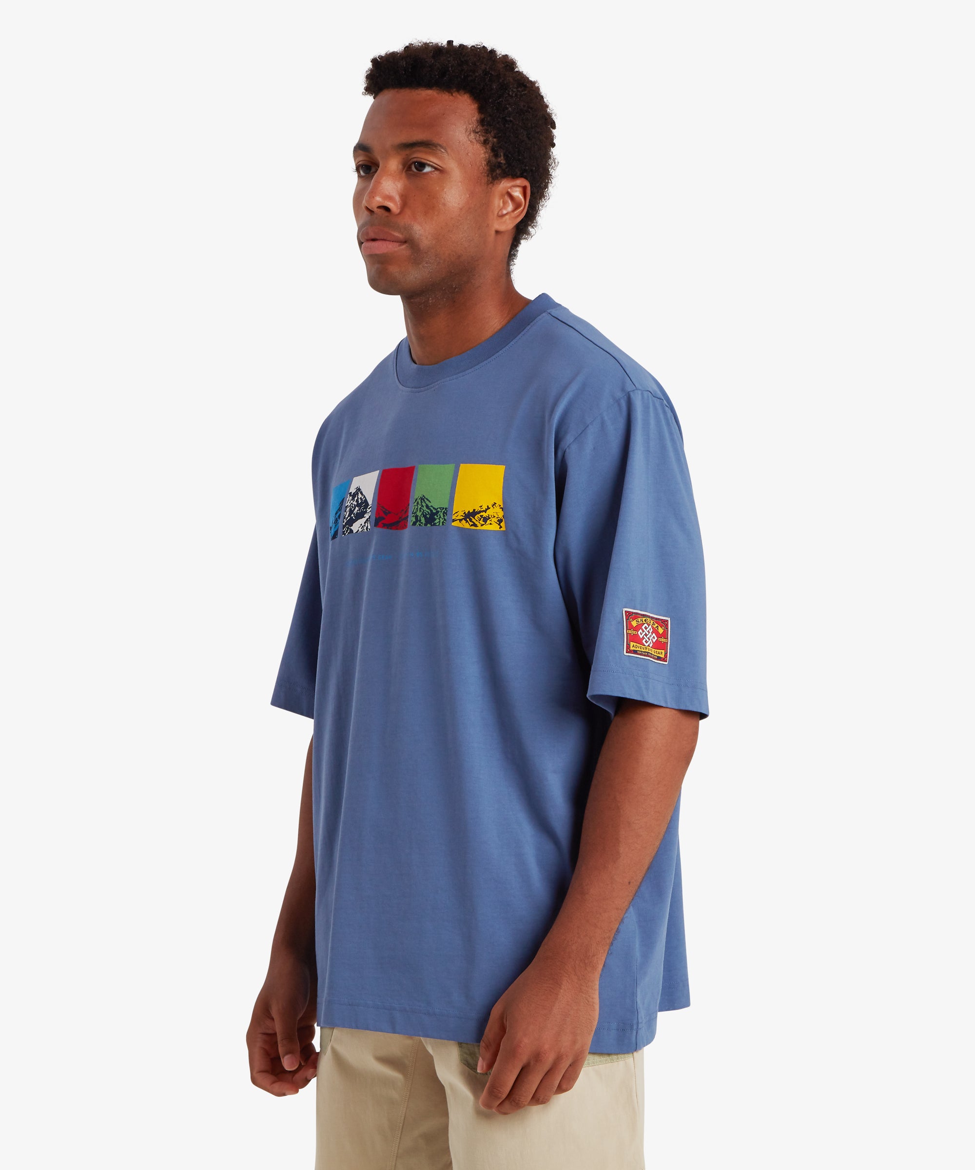 Man wearing a relaxed fit blue T-shirt. The T-shirt features a horizontal graphic across the chest made up of five rectangular color blocks in blue, white, red, green, and yellow, each containing a mountain illustration. Beneath the graphic the words "Sherpa Adventure Gear" are printed in a light blue font. On the left sleeve is red, rectangular mountaineering patch.  The outfit is completed with a pair of beige shorts with pale green pockets. The man is standing with is body turned slightly to the right.