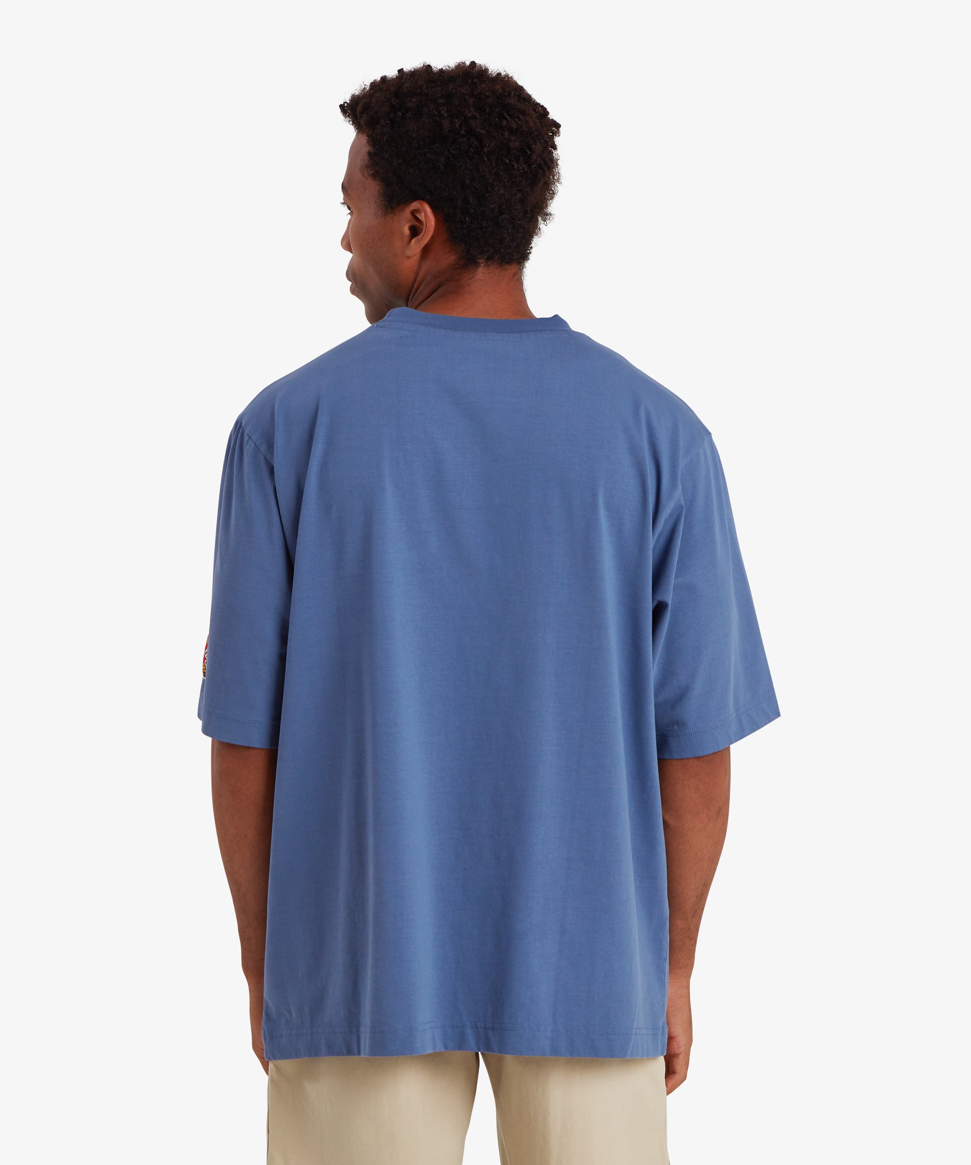 A back view of a man wearing relaxed fit, baggy blue T-shirt. The T-shirt is plain of the man with no graphic or detail. The T-shirt is paired with beige shorts.