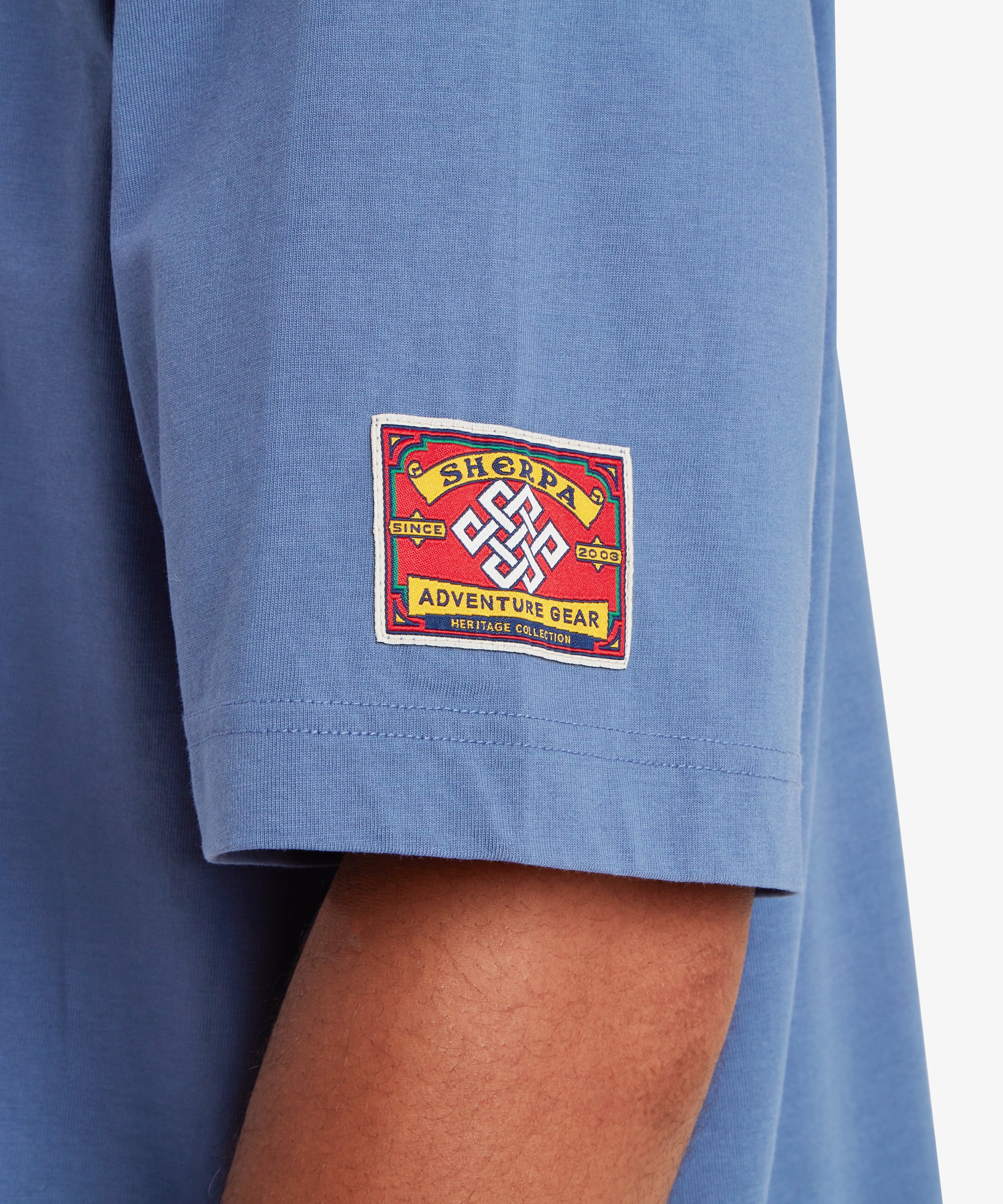 A close-up view of the left sleeve of a blue T-shirt. A square red woven patch is stitched near the sleeve hem, featuring a white endless knot symbol at the center. The patch reads “Sherpa Adventure Gear” with “Since 2003” and “Heritage Collection” incorporated into the design. The fabric appears soft with a relaxed drape, set against a white background.