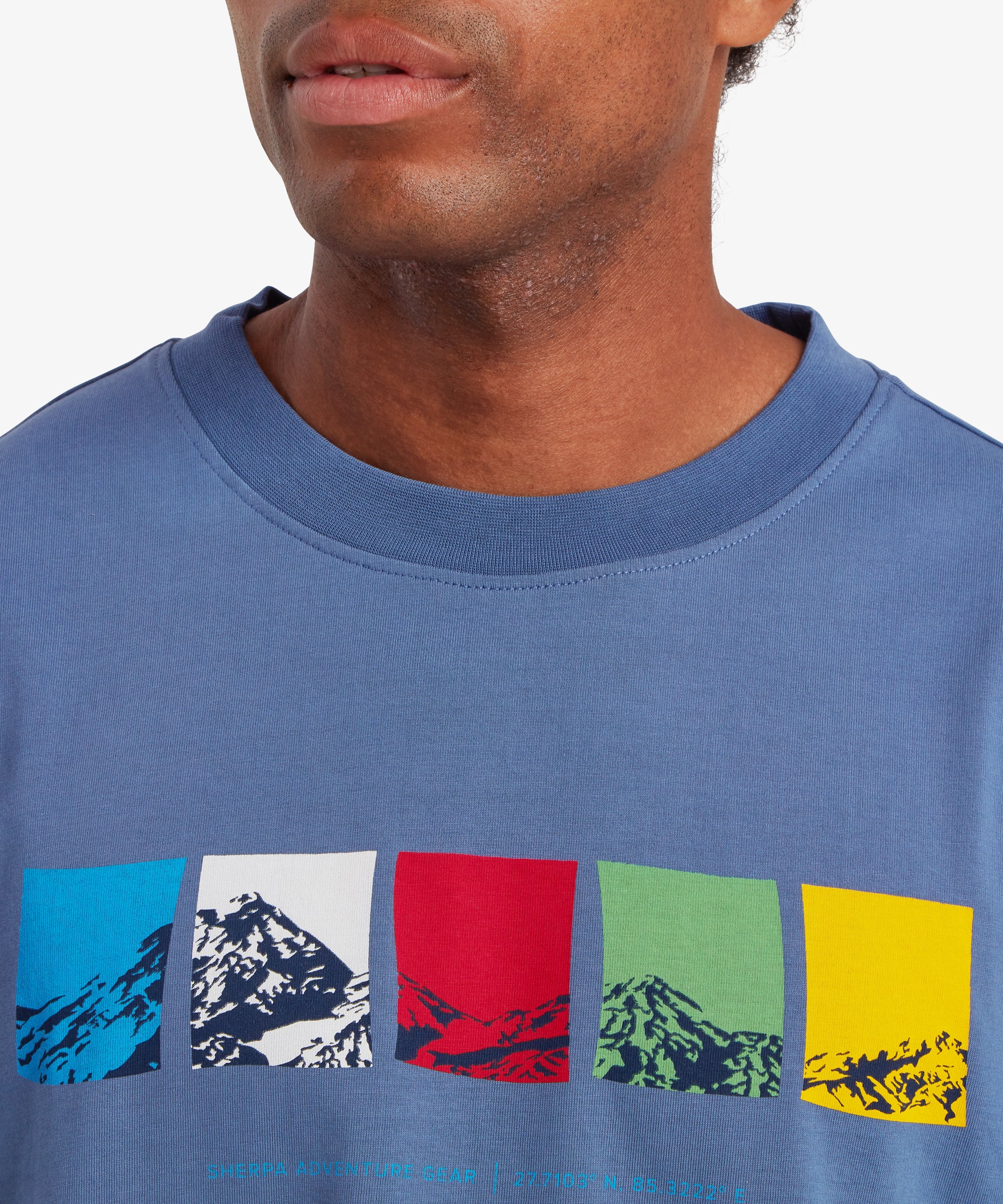 A close-up view of a man wearing a blue T-shirt. The man's upper chest and the neckline of the T-shirt are visible. The T-shirt has a ribbed crew neck collar and features a horizontal graphic across the chest made up of five rectangular color blocks in blue, white, red, green, and yellow, each containing a stylized mountain illustration. Beneath the graphic is small text reading “Sherpa Adventure Gear” followed by geographic coordinates. The man is standing against a white background.