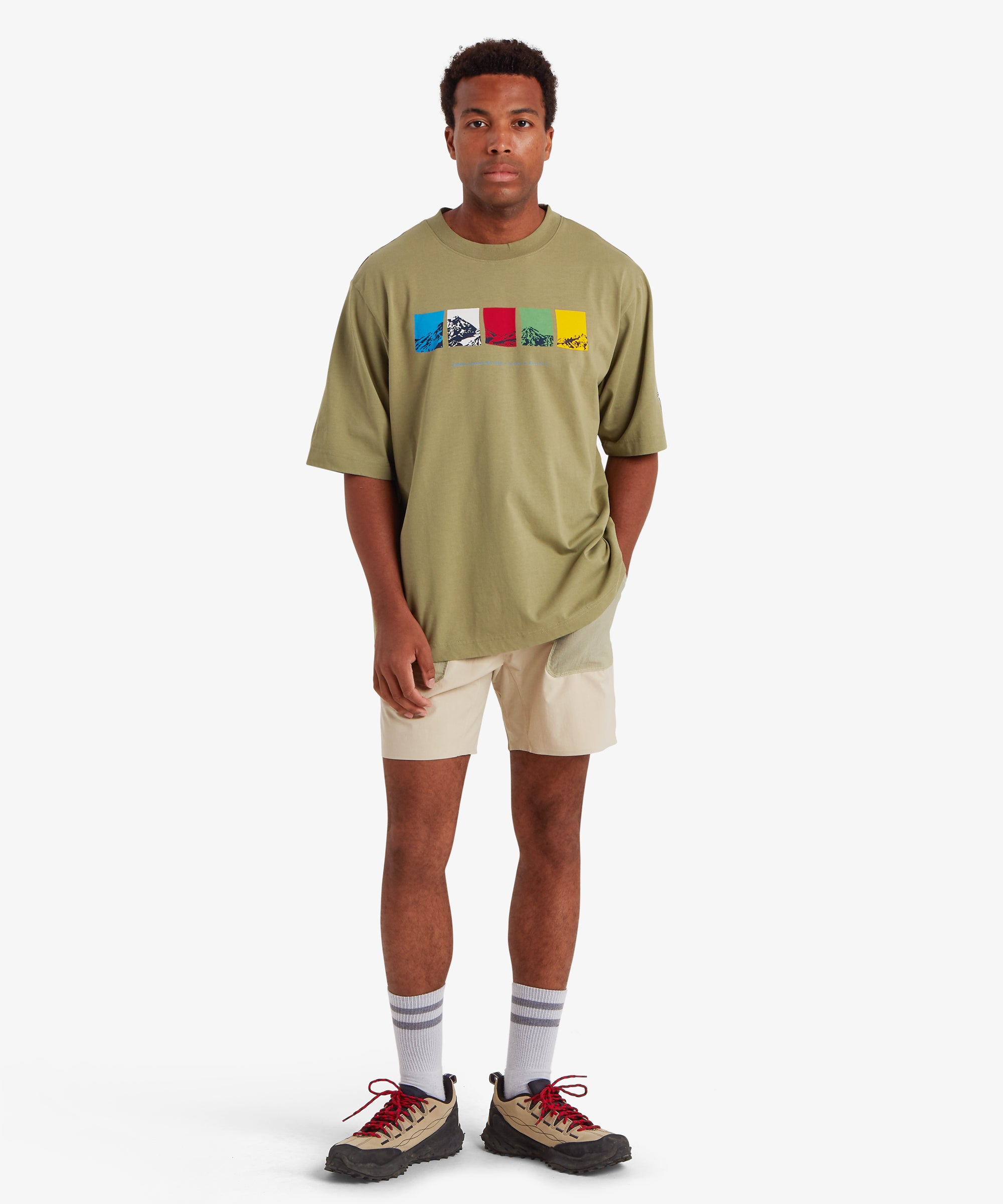 Man wearing a loose fitting green T-shirt. The T-shirt features a horizontal graphic across the chest made up of five rectangular color blocks in blue, white, red, green, and yellow, each containing a mountain illustration. Below the graphic the words "Sherpa Adventure Gear" are printed in a light blue font. On the left sleeve is red, rectangular mountaineering patch. The outfit is styled with of beige shorts, white socks and beige walking shoes with red laces. The man has his head slightly to the left.