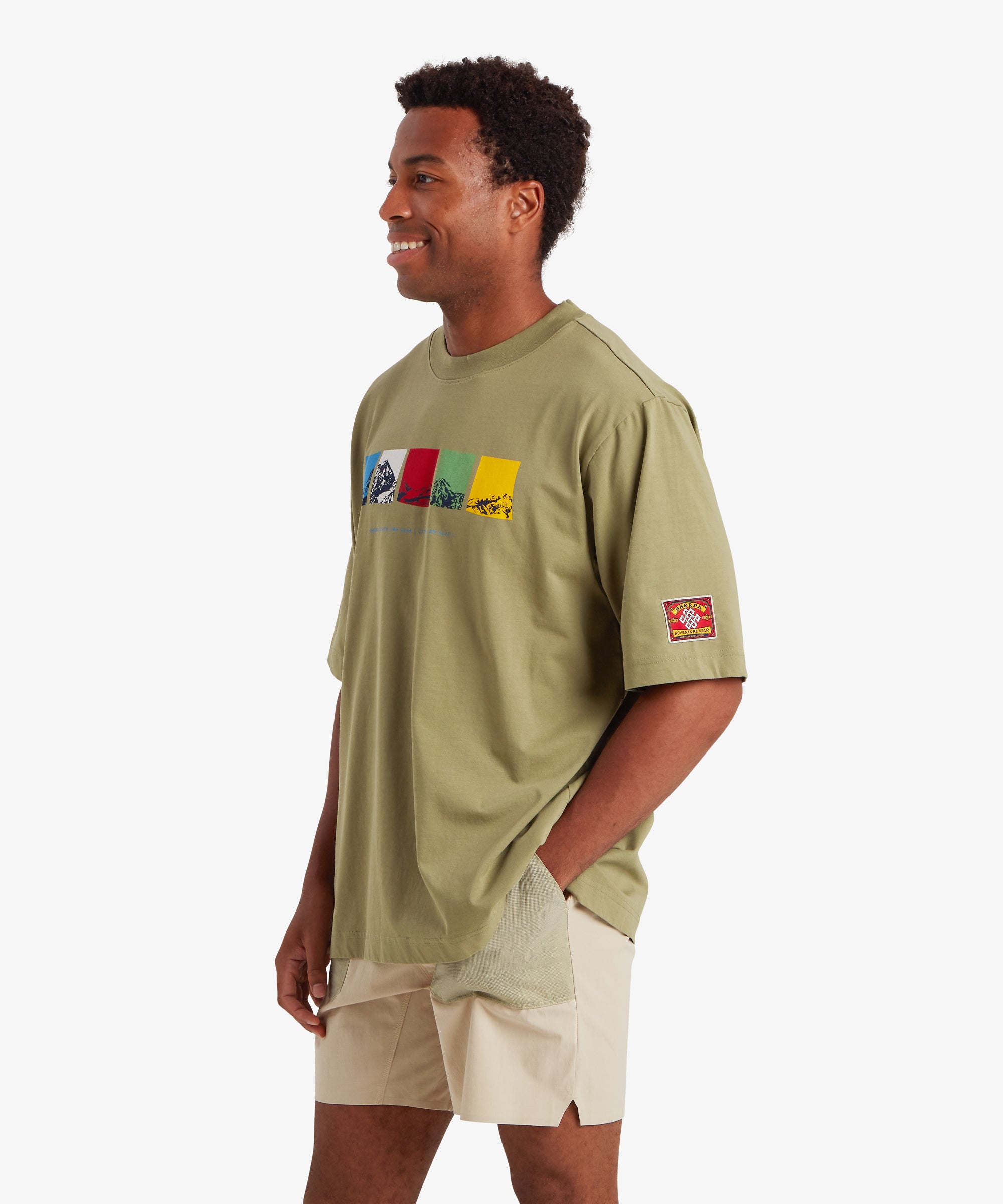 Man wearing a relaxed fit green T-shirt. The T-shirt features a horizontal graphic across the chest made up of five rectangular color blocks in blue, white, red, green, and yellow, each containing a mountain illustration. Beneath the graphic the words "Sherpa Adventure Gear" are printed in a light blue font. On the left sleeve is red, rectangular mountaineering patch.  The outfit is completed with a pair of beige shorts with pale green pockets. The man is standing with is body turned slightly to the right.