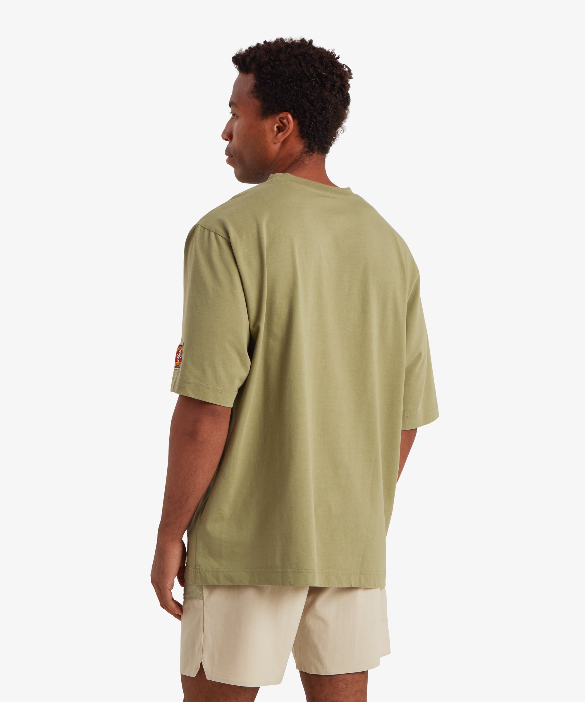 A back view of a man wearing relaxed fit, baggy green T-shirt. The back of T-shirt is plain of the man with no graphic or detail. On the left sleeve is a rectangular mountaineering patch. At the centre of the patch is a white endless knot graphic. The words "Sherpa adventure Gear" are stitched above and below the endless knot. The T-shirt is paired with beige above the knee shorts.