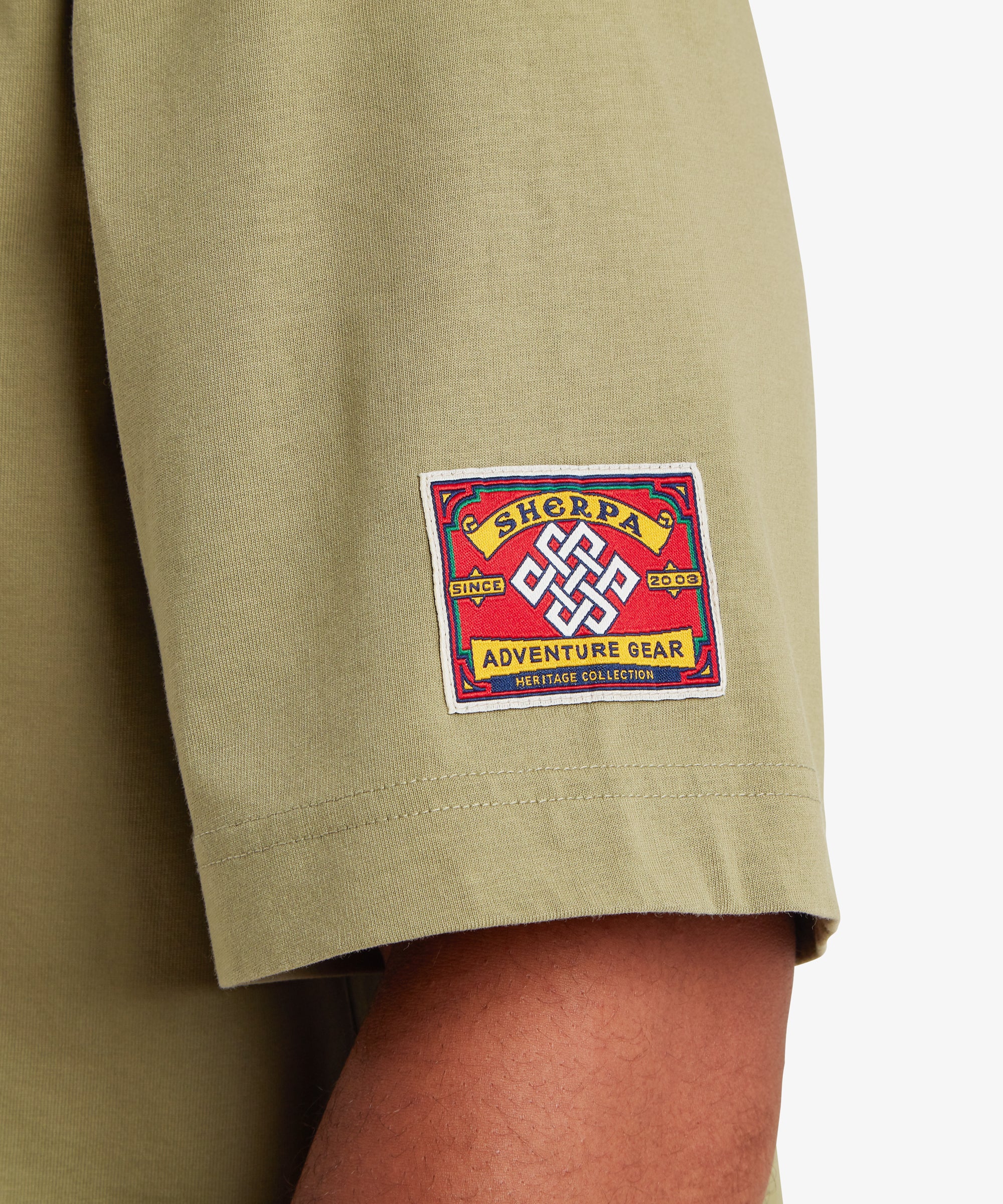A close-up view of the left sleeve of a green T-shirt. A square red woven patch is stitched near the sleeve hem, featuring a white endless knot symbol at the center. The patch reads “Sherpa Adventure Gear” with “Since 2003” and “Heritage Collection” incorporated into the design. The fabric appears soft with a relaxed drape, set against a white background.
