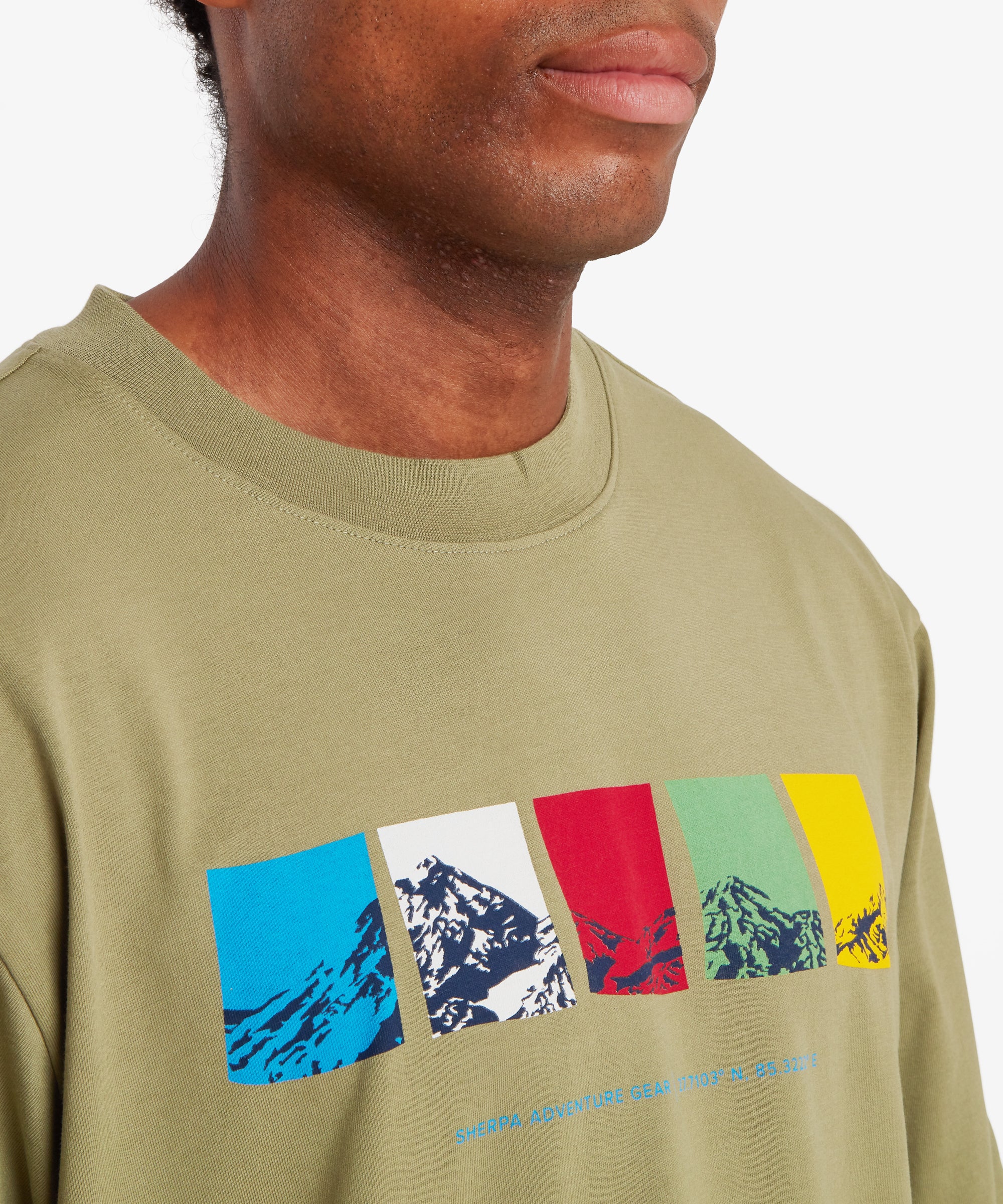 A close-up view of a man wearing a green T-shirt. The man's upper chest and the neckline of the T-shirt are visible. The T-shirt has a ribbed crew neck collar and features a horizontal graphic across the chest made up of five rectangular color blocks in blue, white, red, green, and yellow, each containing a stylized mountain illustration. Beneath the graphic is small text reading “Sherpa Adventure Gear” followed by geographic coordinates. The man is standing against a white background.