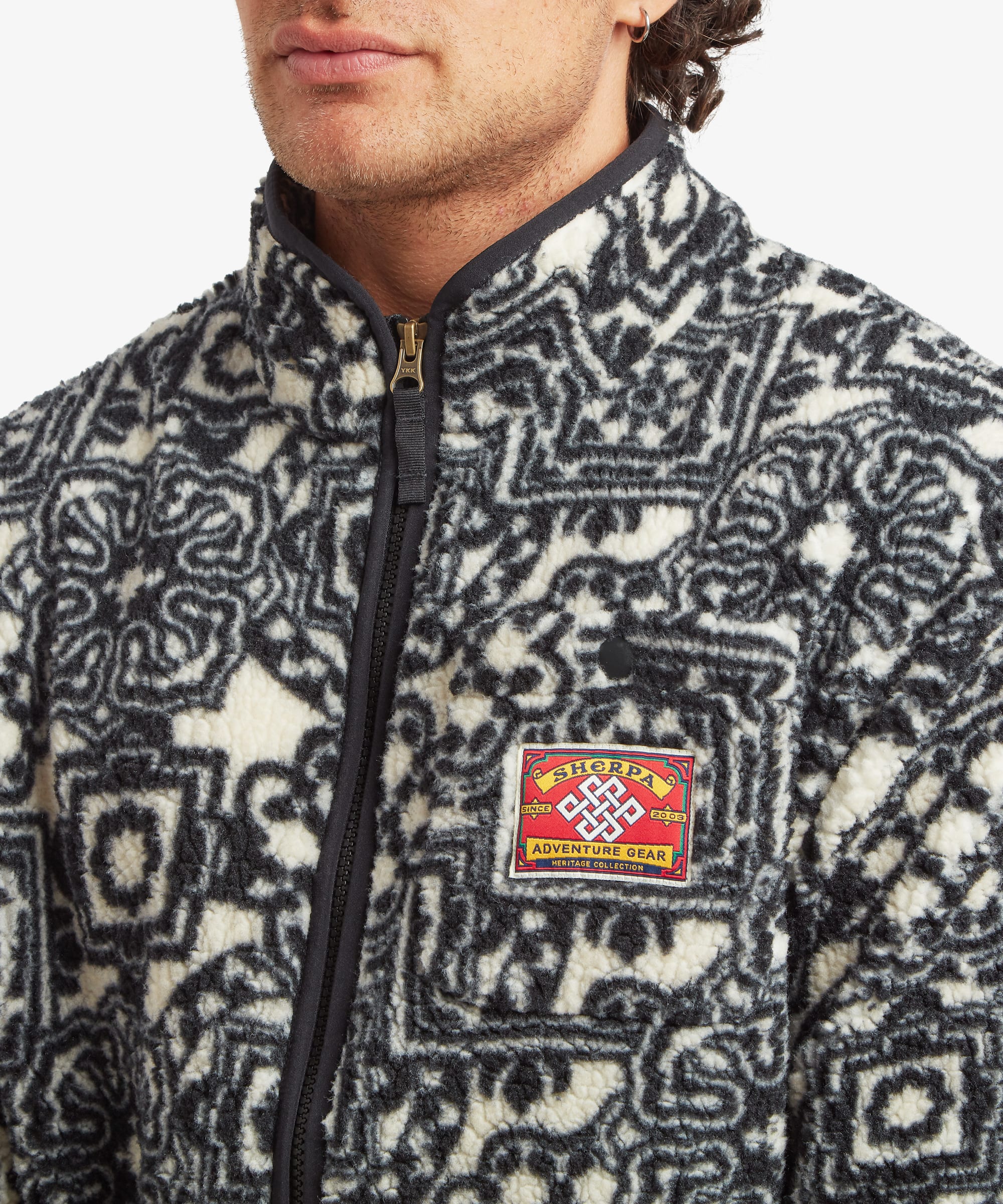 Close-up of a black printed full-zip fleece jacket featuring an allover geometric pattern, stand collar, and contrast chest patch.
