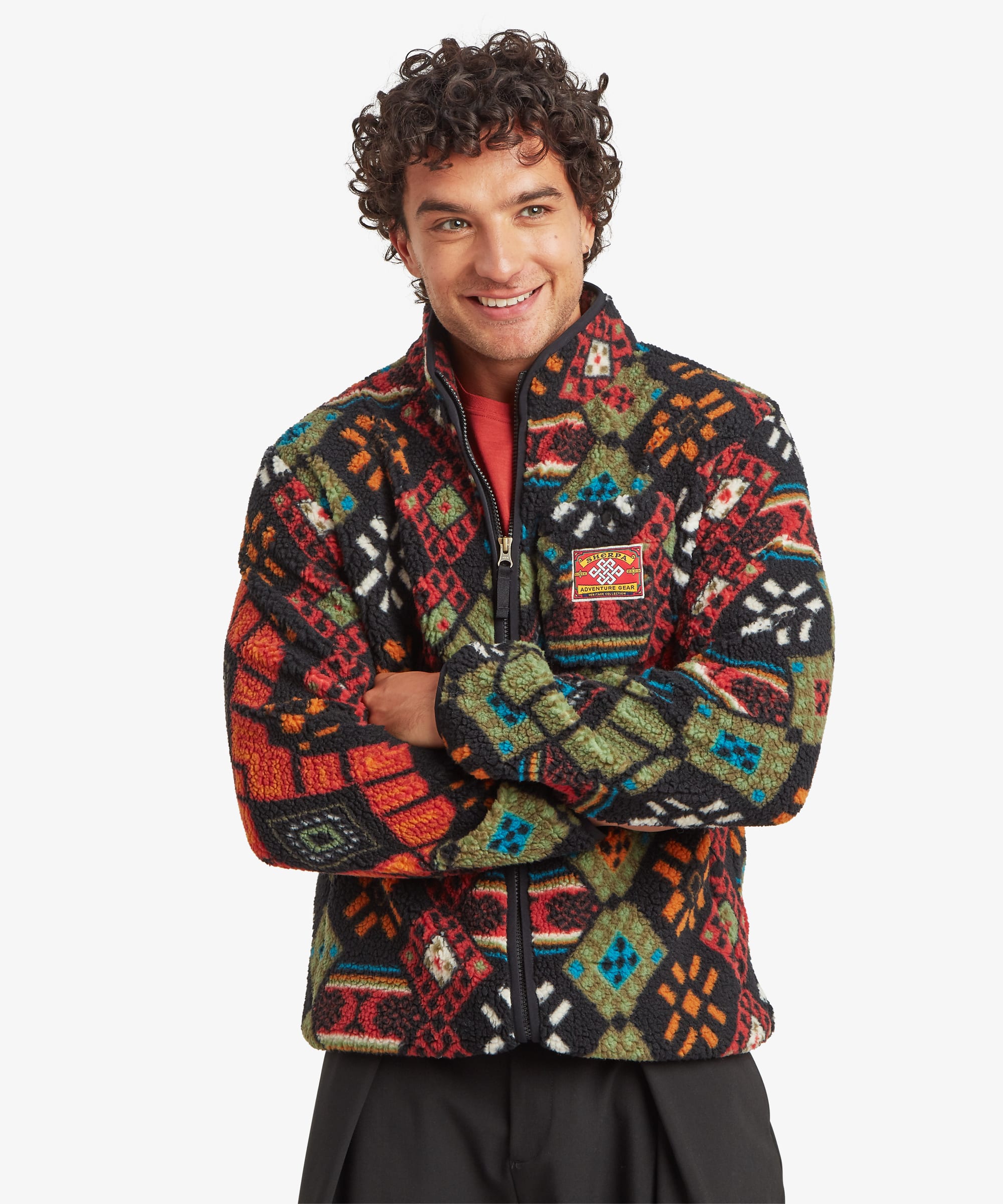 Front view of a smiling male model wearing the Sherpa Tarcho Printed Full Zip Jacket in black with a multicoloured geometric pattern and black trousers. The fleece jacket features a high stand collar, full-length front zip, elasticated cuffs and hem, a chest patch with the Sherpa logo, and a relaxed fit.