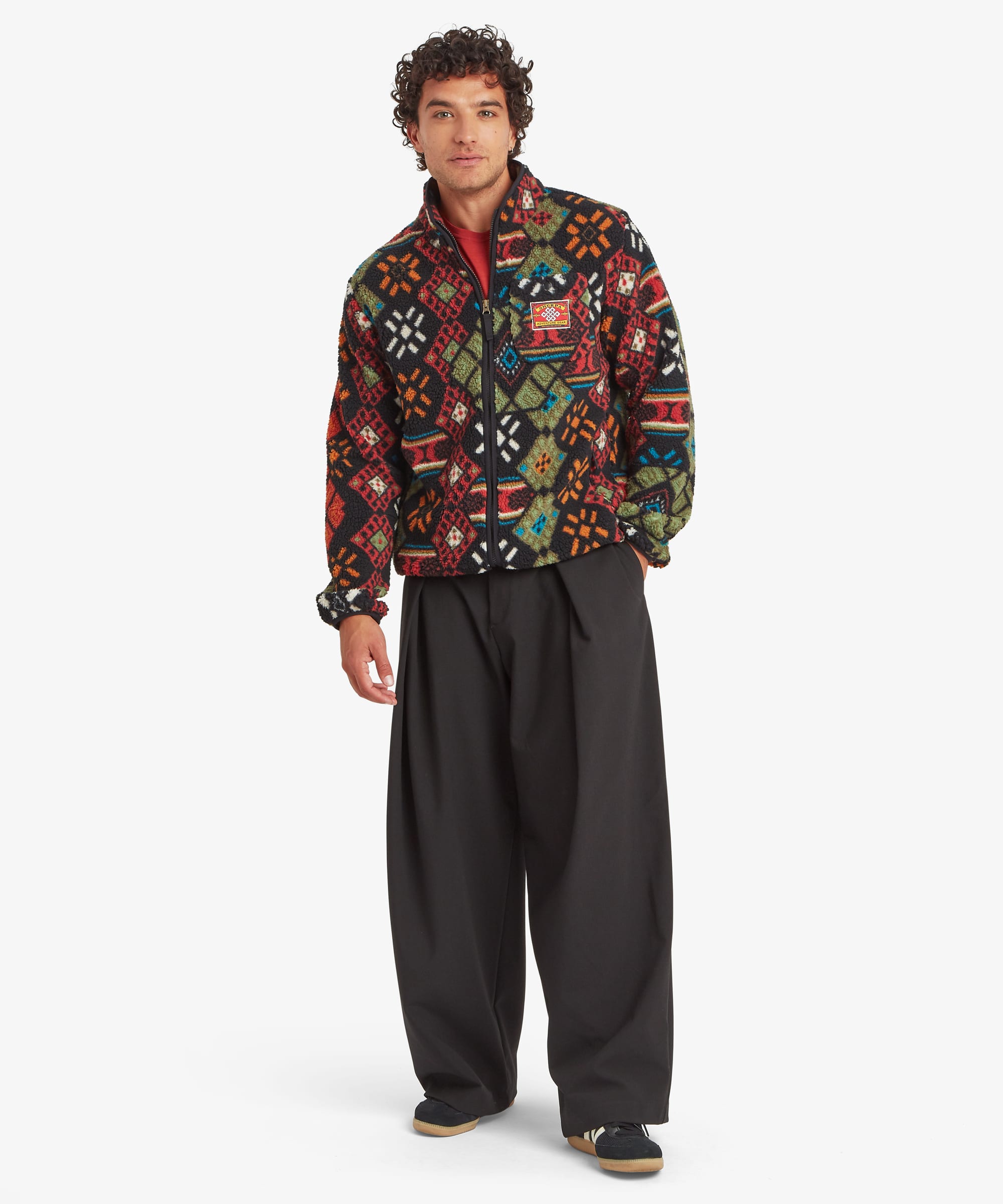 Full-length front view of a male model wearing the Sherpa Tarcho Printed Full Zip Jacket in black with a vibrant red, green, blue and orange geometric print. The textured fleece jacket has a stand collar, full zip fastening, long sleeves, side pockets and a relaxed outdoor fit.