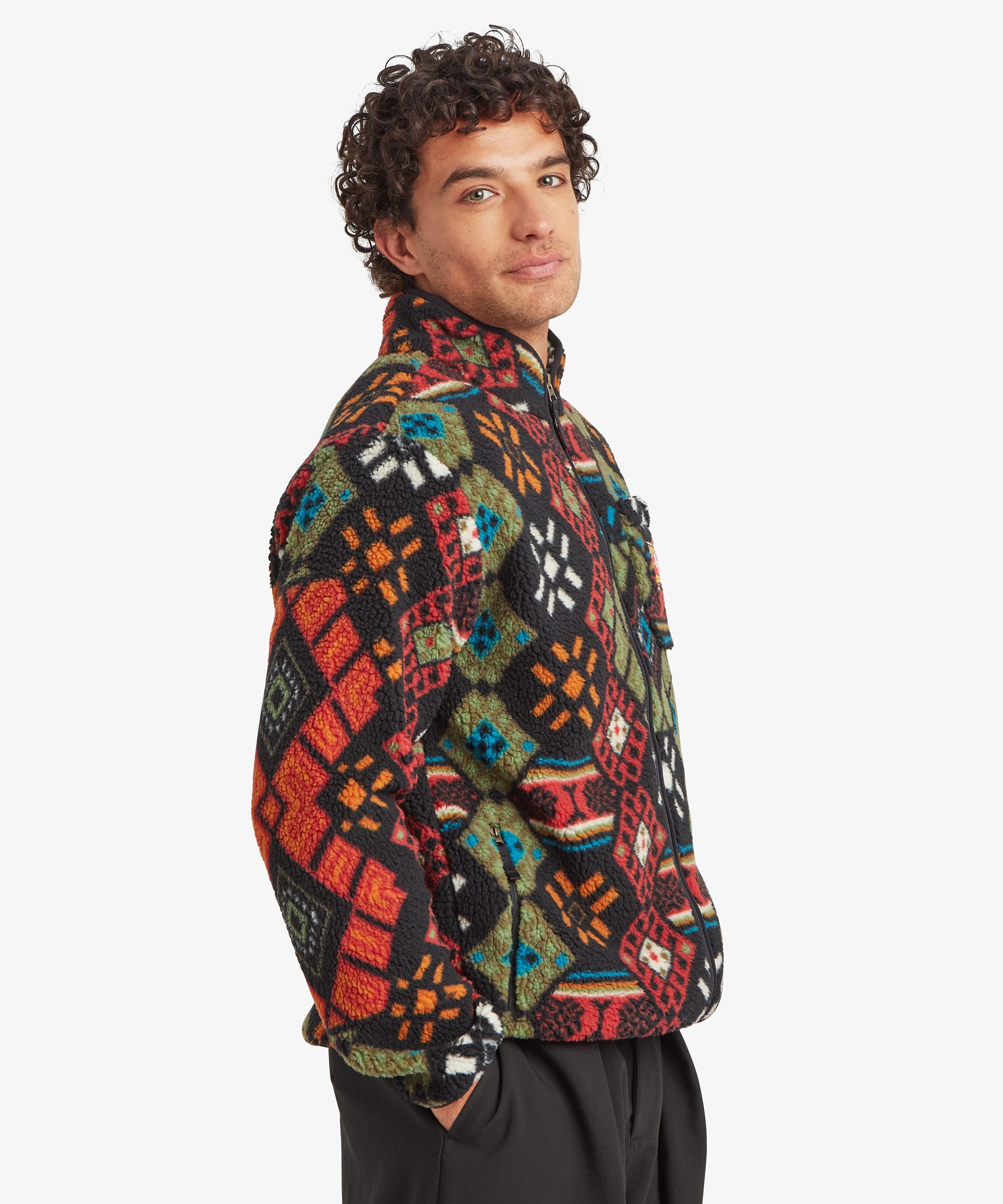 Side profile of a male model wearing the Sherpa Tarcho Printed Full Zip Jacket in black with a colourful all-over geometric pattern. The image highlights the high collar, relaxed fit, side pocket and the soft fleece texture of the jacket.