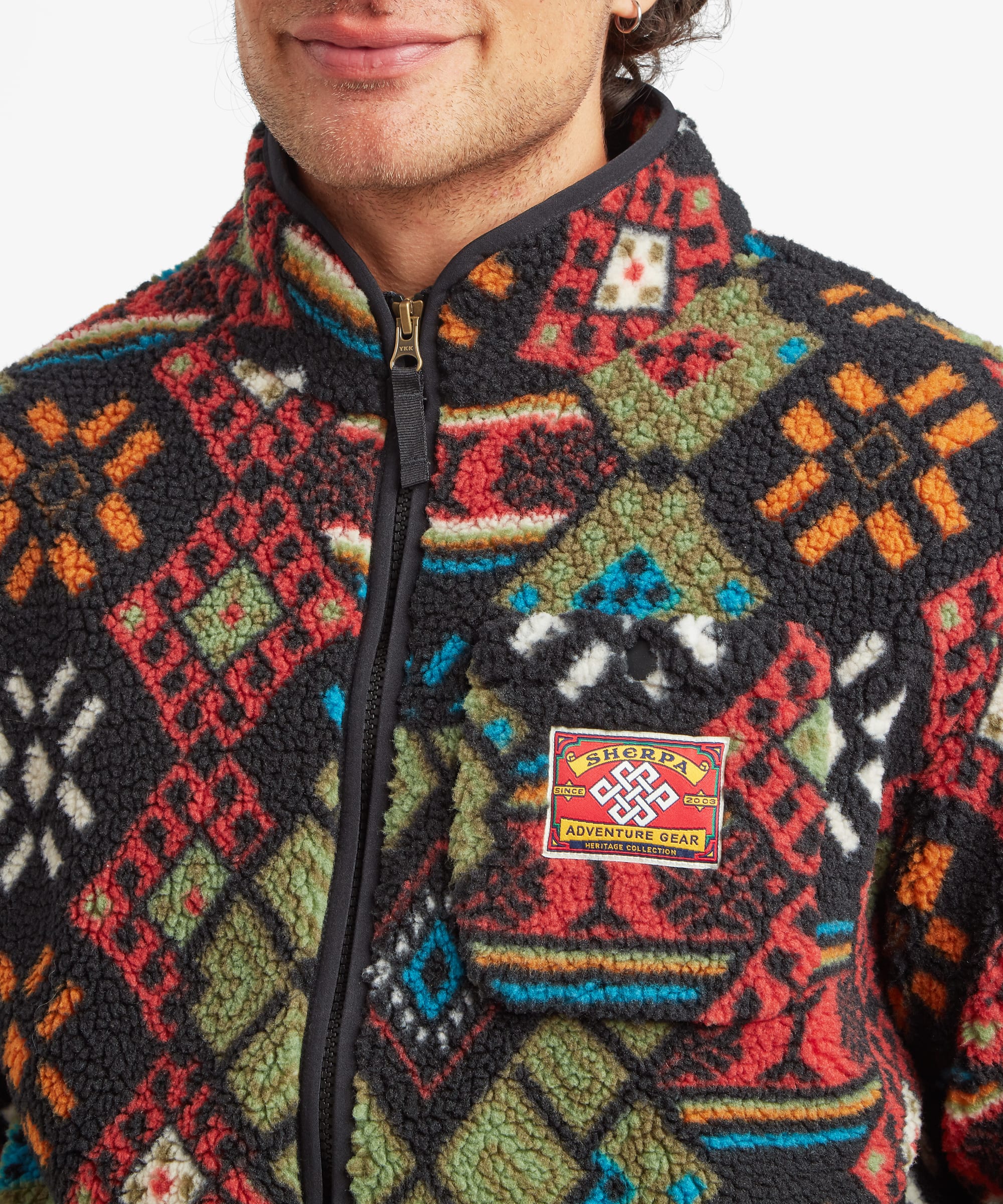 Close-up of a male model wearing the Sherpa Tarcho Printed Full Zip Jacket in black, highlighting the textured fleece fabric, stand collar, full front zip and woven Sherpa logo patch on the chest.