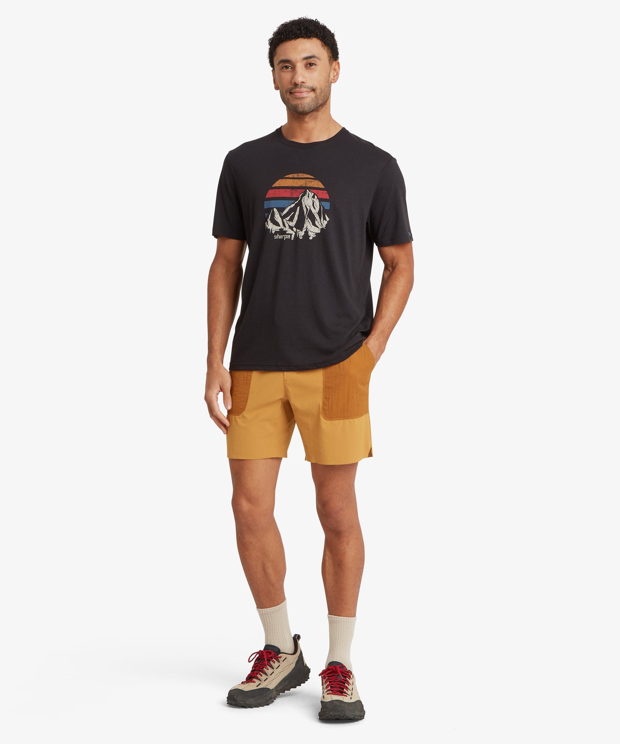 A full-length view of a person wearing mustard-orange outdoor shorts with contrasting darker orange ripstop patch pockets on the front. The shorts have an elastic waistband with a yellow drawcord. Styled with a black graphic T-shirt featuring a mountain design and neutral hiking shoes with red laces.