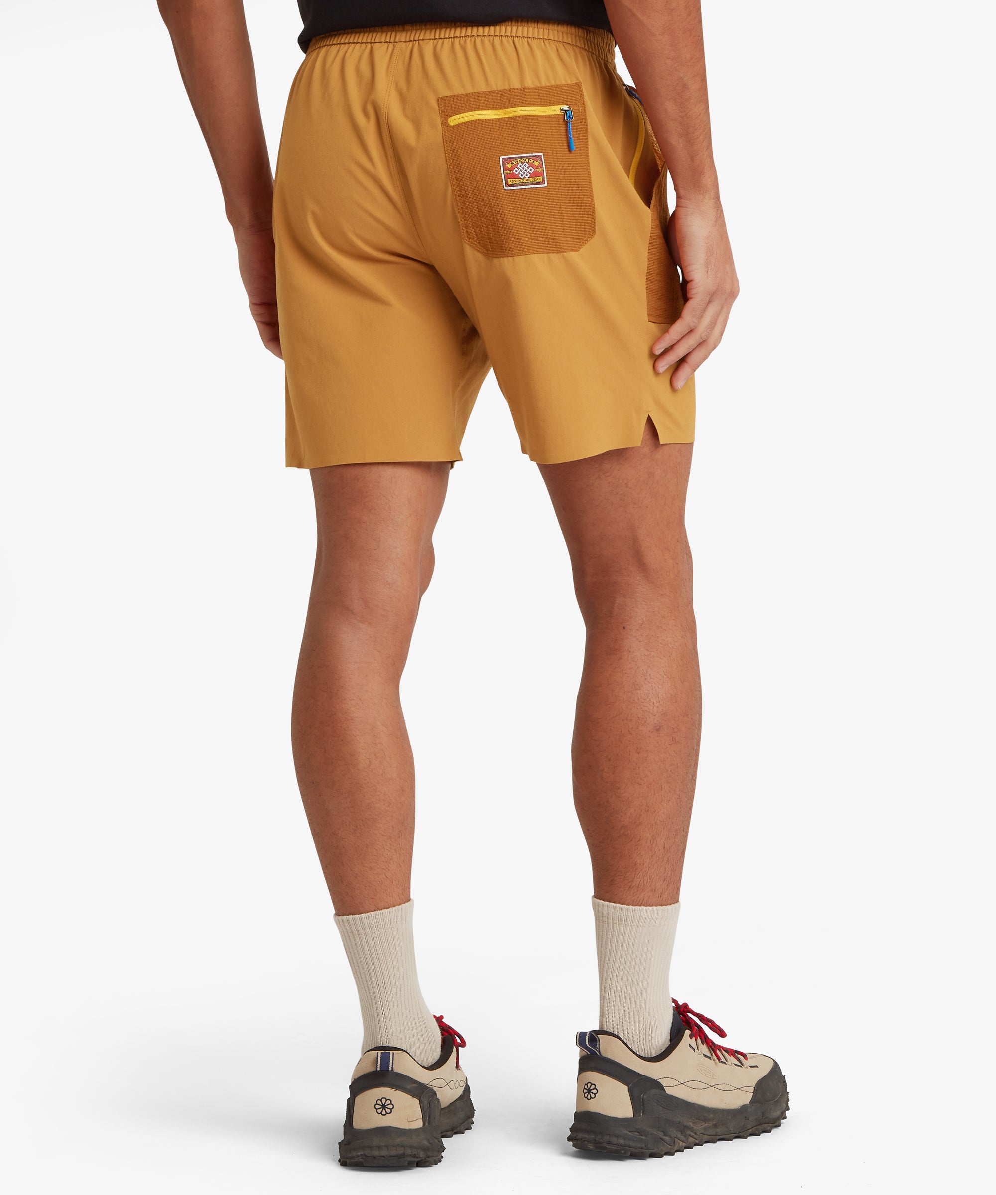 A rear view of mustard-orange performance shorts showing a contrasting darker orange ripstop back pocket with a bright yellow zipper and woven adventure‑gear patch, plus small side vents at the hem.