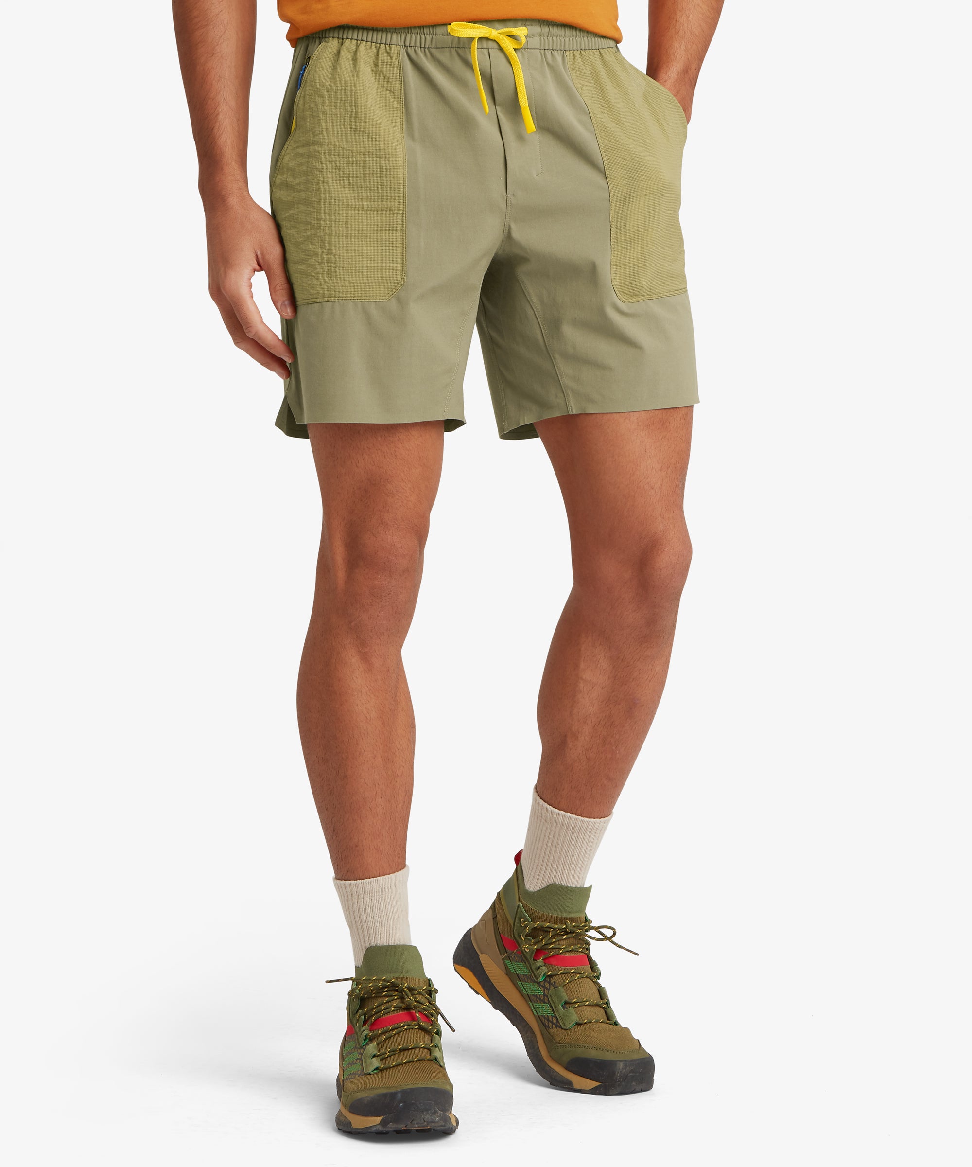 A front view of light olive outdoor shorts with contrasting olive ripstop patch pockets and a bright yellow drawstring. Styled with outdoor trainers and a short-sleeve top.