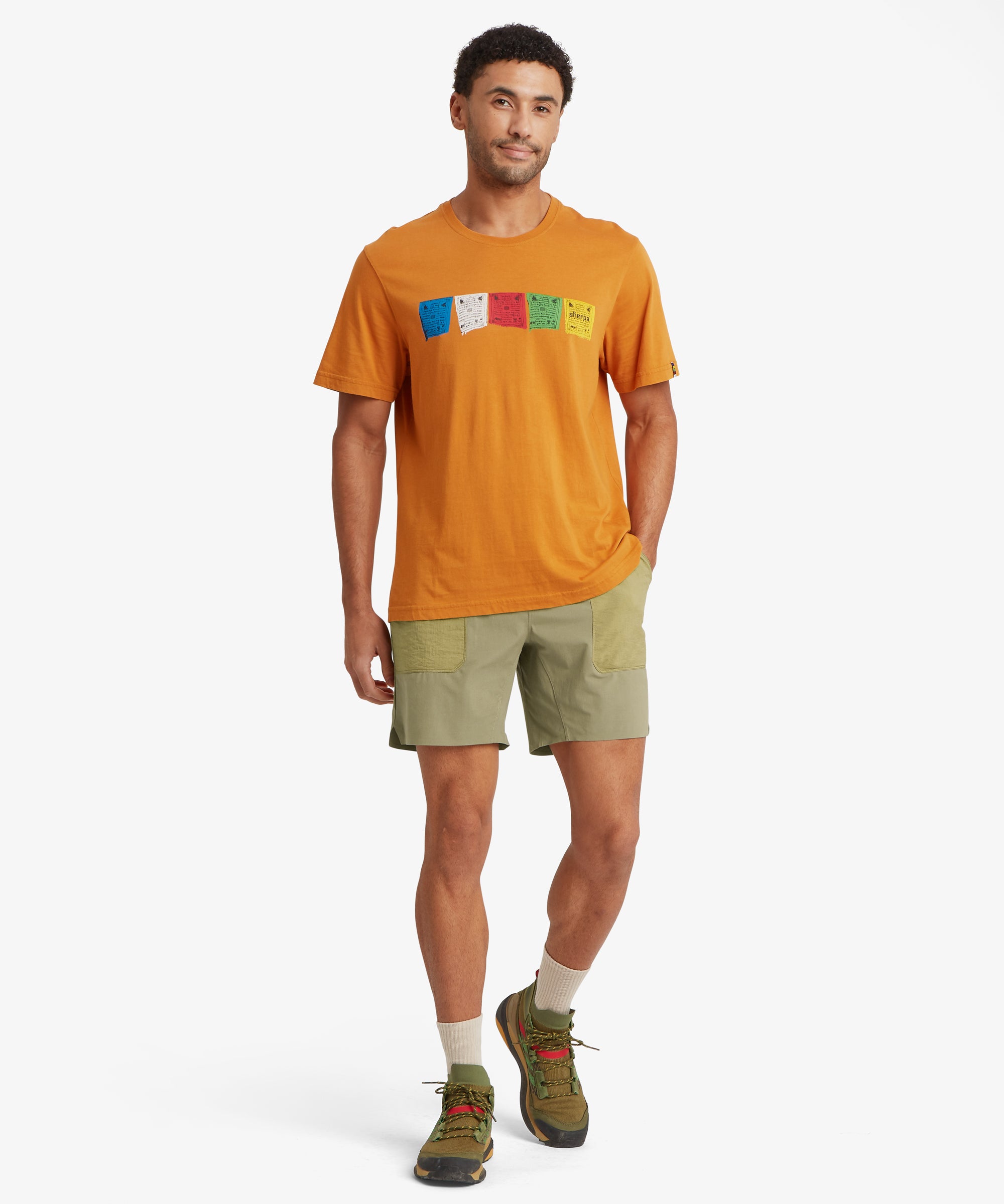 A full-length view of light olive shorts featuring oversized ripstop patch pockets and a relaxed elastic waistband. Paired with an orange T-shirt printed with multicolored flag graphics and green hiking shoes.