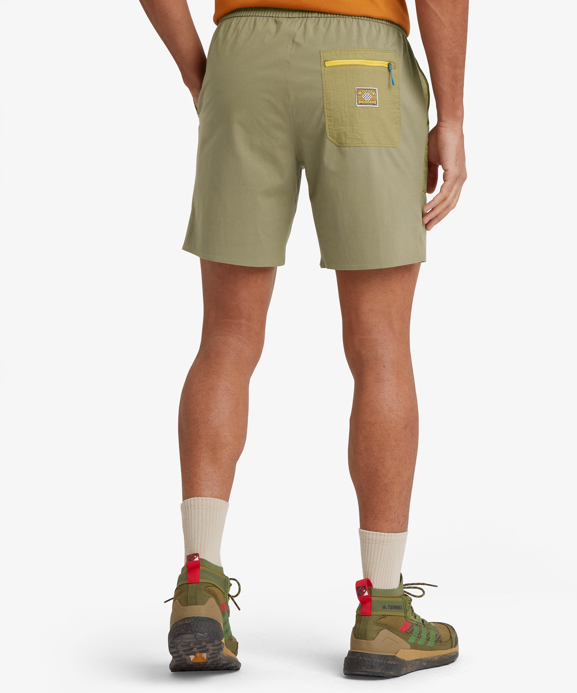 A rear view of light olive shorts with a contrasting darker olive ripstop back pocket and bright yellow zipper, plus small side hem vents and outdoor footwear.
