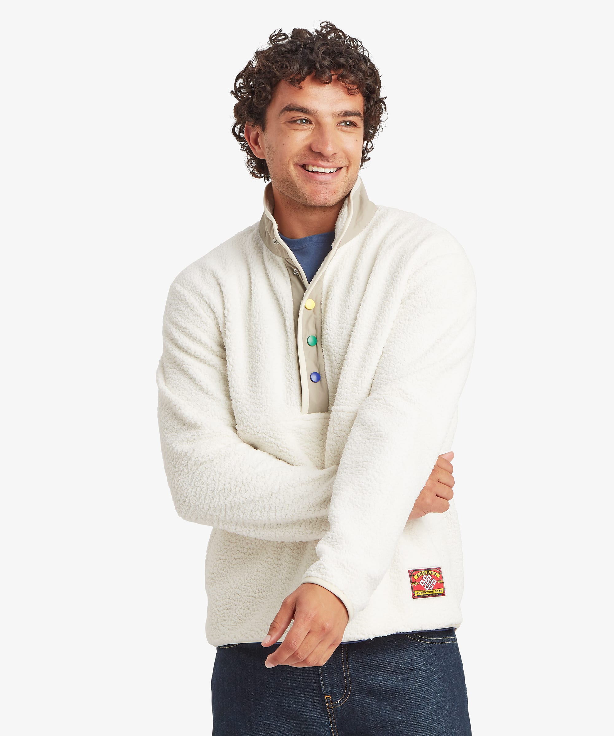 Front view of a male model wearing the Sherpa Tarcho Snap Pullover in white with dark blue jeans. The textured fleece pullover features a high stand collar, multicoloured snap-button placket, contrast trim, long sleeves, hand pockets and a relaxed fit.
