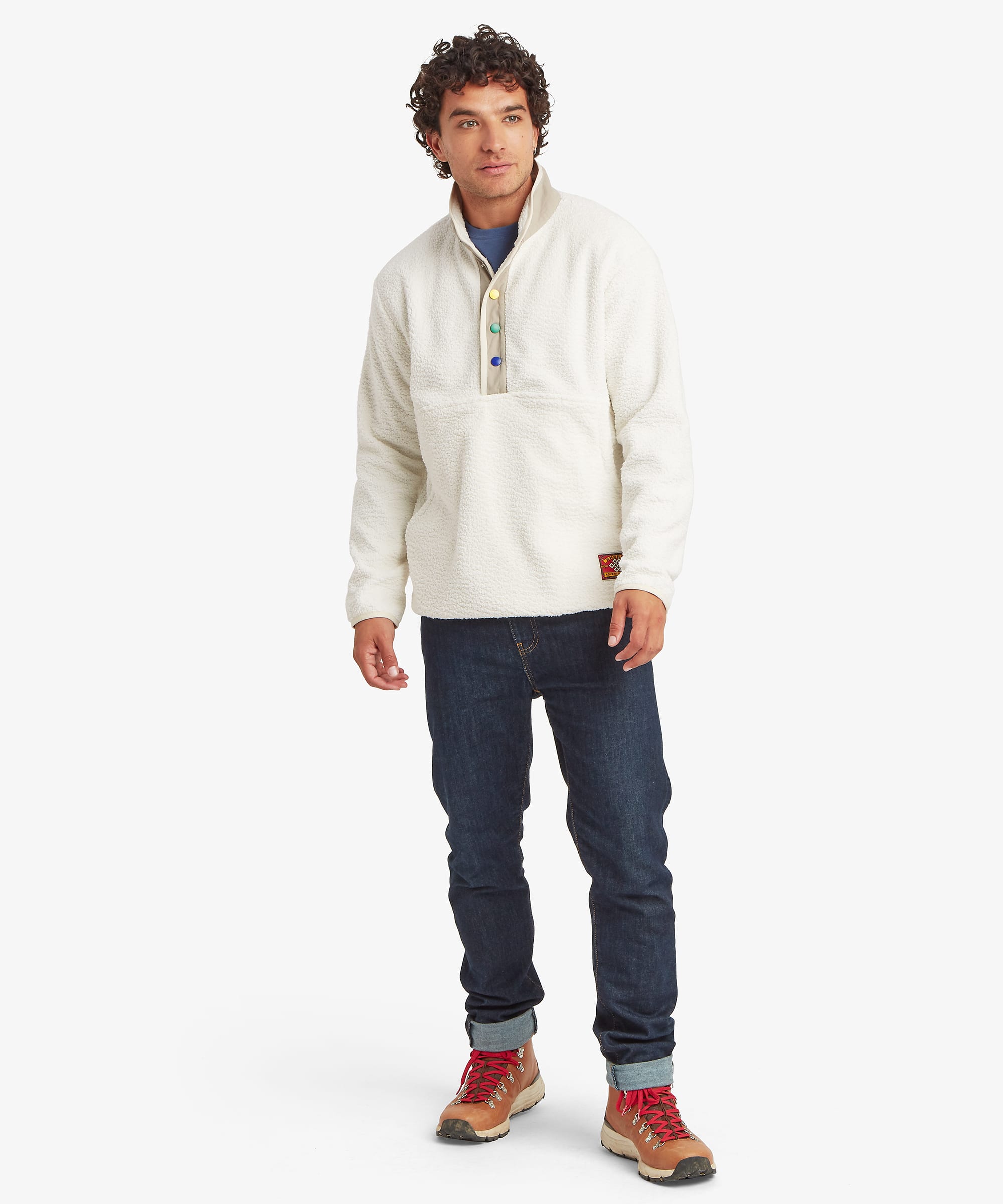 Full-length front view of a male model wearing the Sherpa Tarcho Snap Pullover in white with dark blue jeans, highlighting the relaxed fit, textured fleece fabric, stand collar, colourful snap placket and hand pockets.