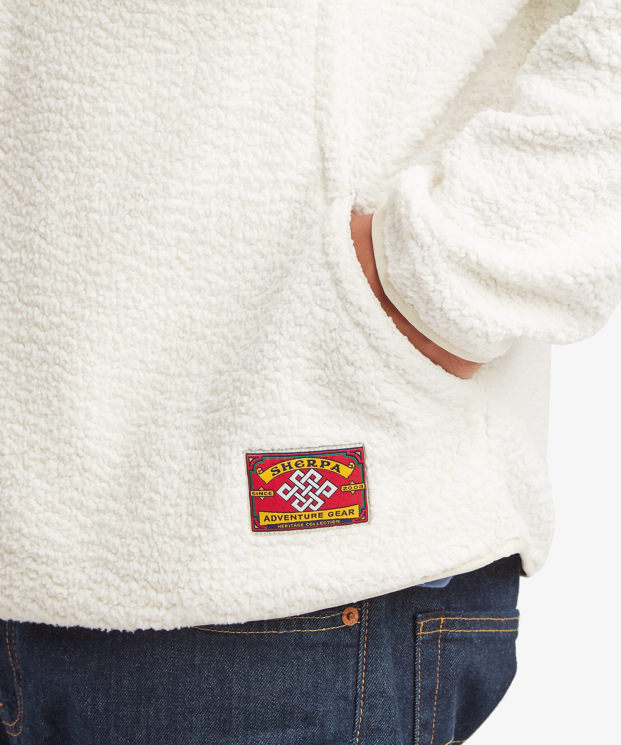 Close-up of the Sherpa Tarcho Snap Pullover in white showing the textured fleece fabric, hand pocket opening and woven Sherpa logo label near the hem.